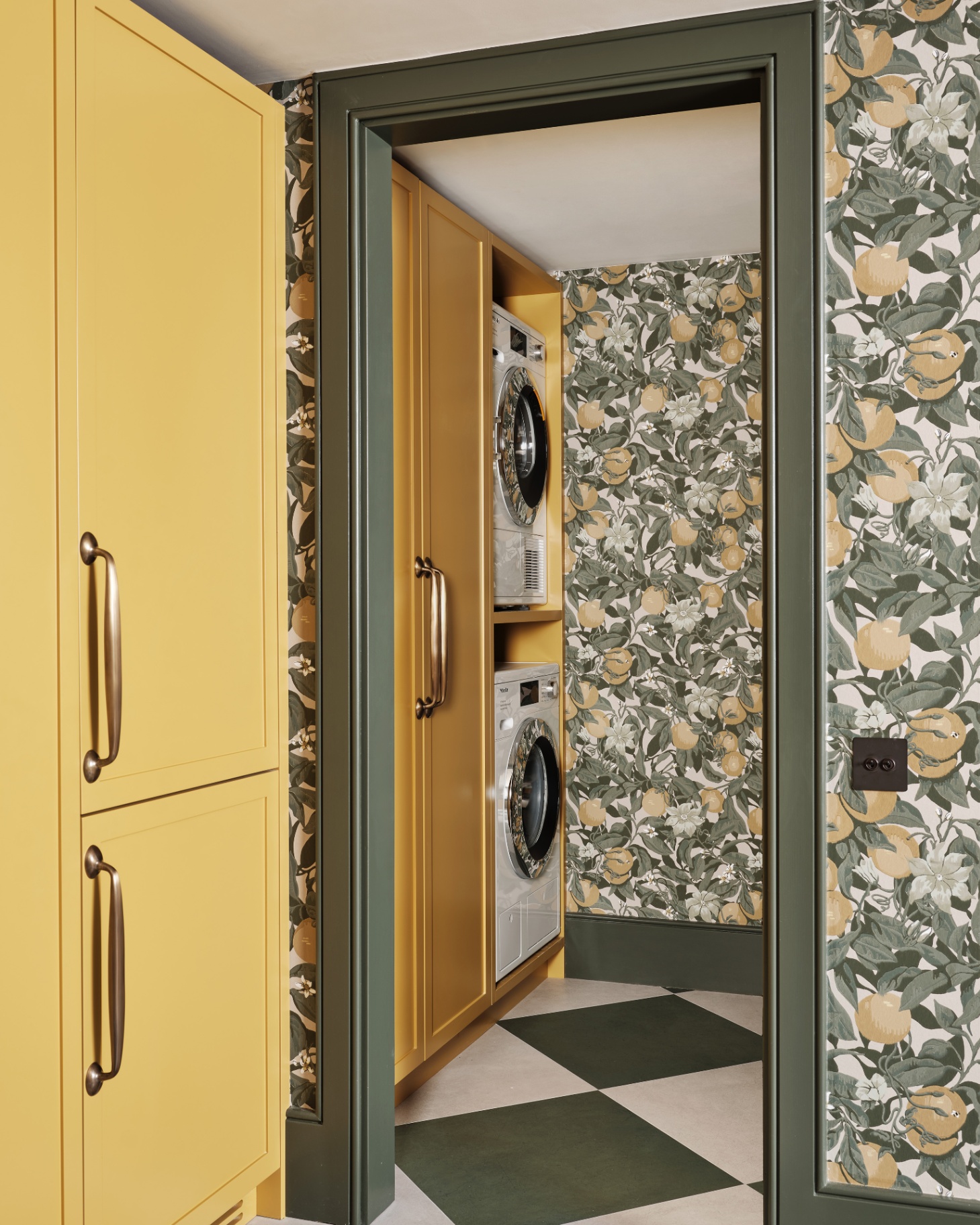 a utility room with two washing machines stacked on top of eachother, green and yellow and white botanical wallpaper, yellow cabinetry and a green painted door frame
