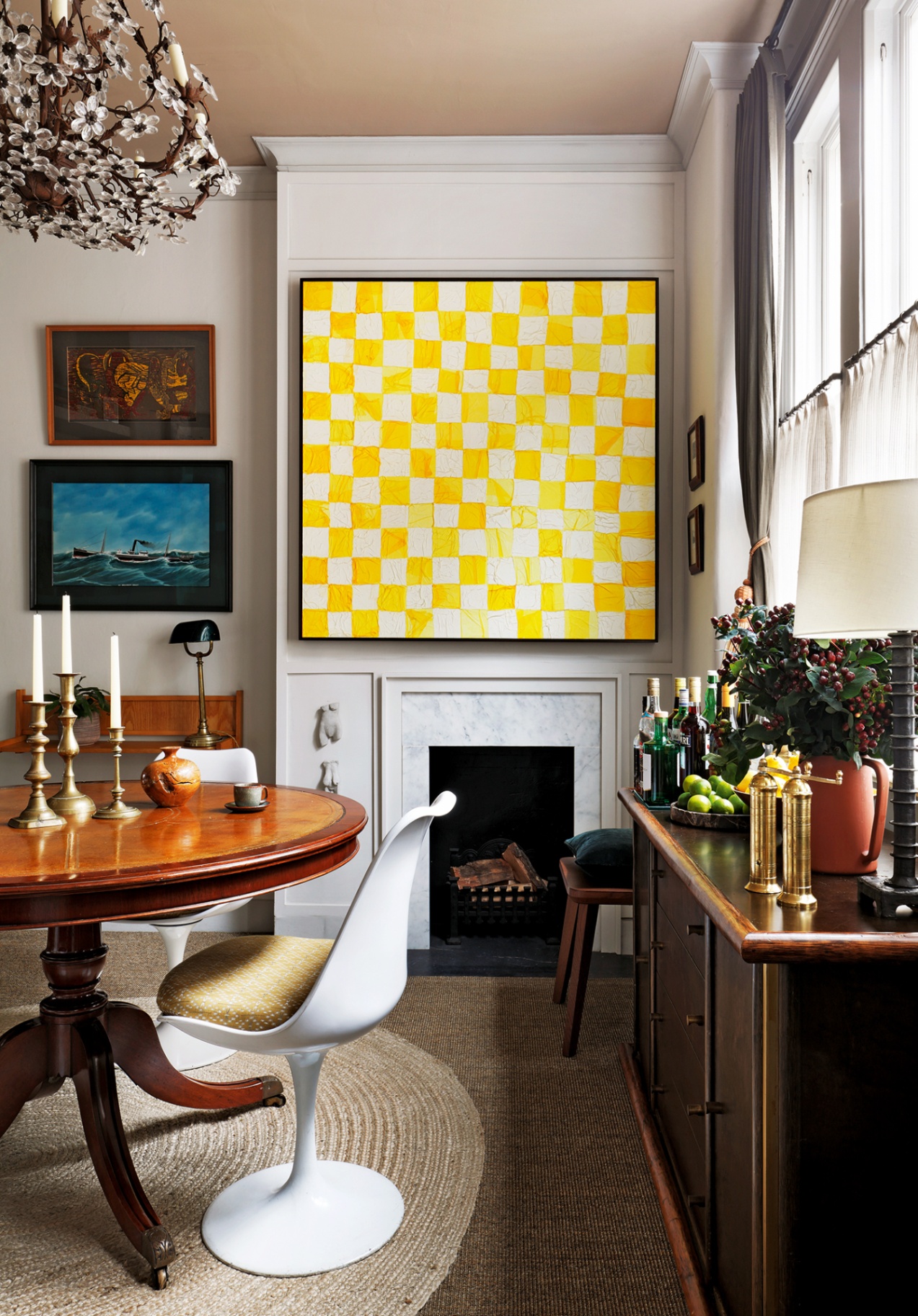 A dining room with a circular wood table and tulip chairs, a wooden sideboard topped with bottles and home accessories. In the background a small fireplace sits beneath a large yellow and white check painting. A floral chandelier hangs above the table