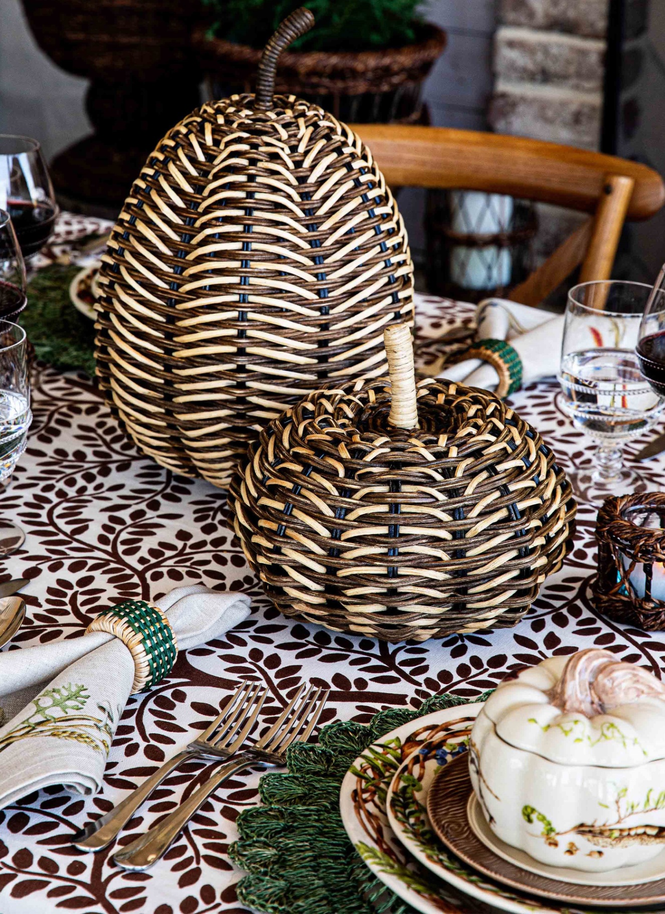 a close up of an autumn table with two large beige and brown rattan pumpkins, a white and brown leaf print tablecloth, clear glasses, a white napkin witha beige and green raffia nakin ring and a stack of plates