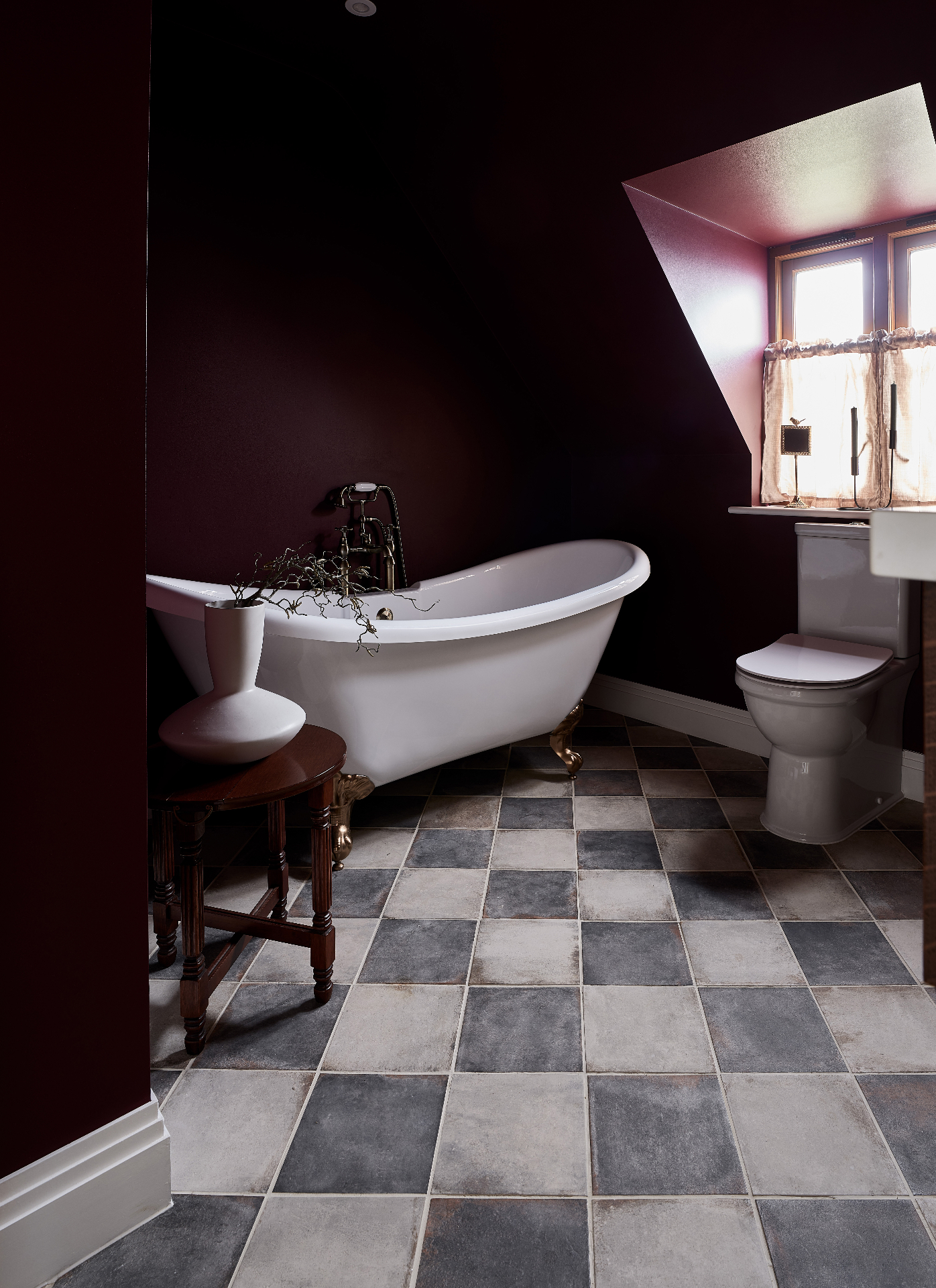 a bathroom with black and white checkerboard floor tiles, a freestanding tub, dark red walls and a small side table with a vase