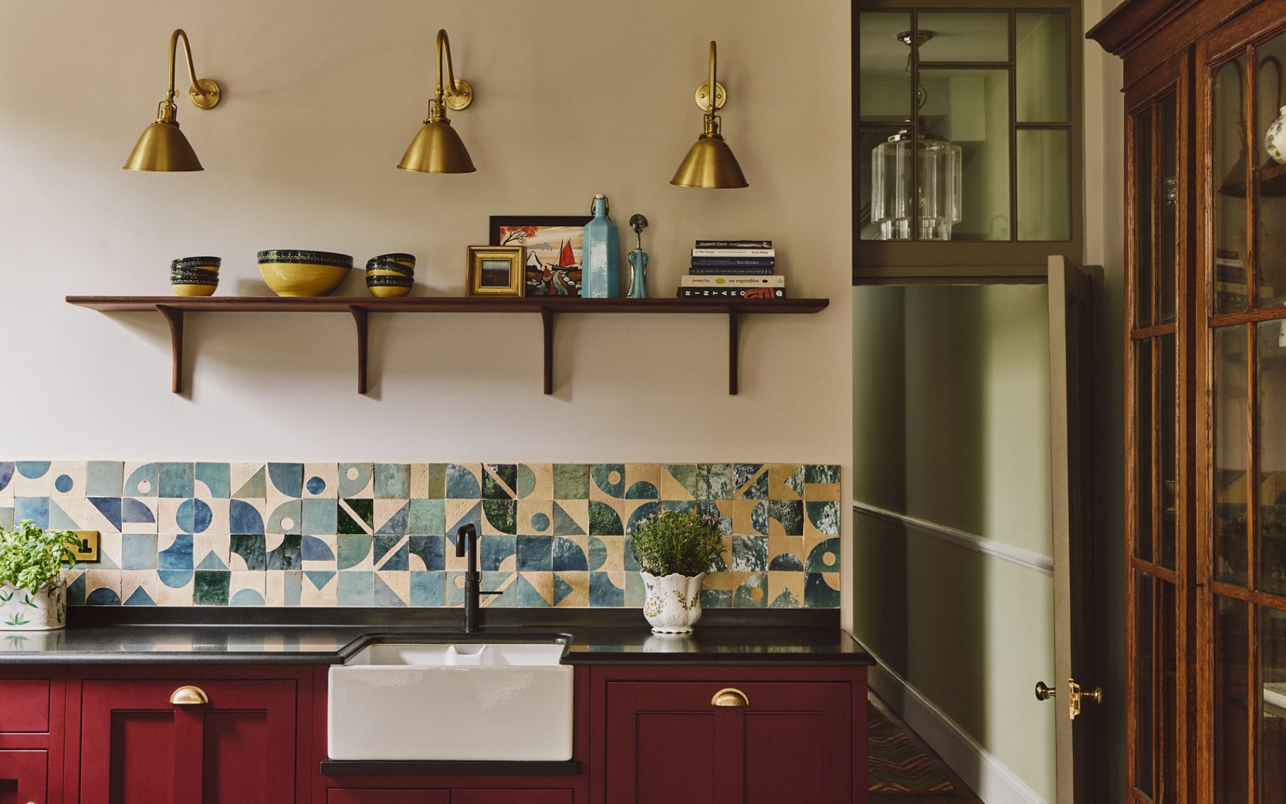 A close up of a kitchen with neutral walls, red cabinets and a tiled blacksplash in blue and white tones with a graphic pattern. The worktops are black granite and a belfast sink is paired with a black tap. Above is a shelf and three brass wall lights
