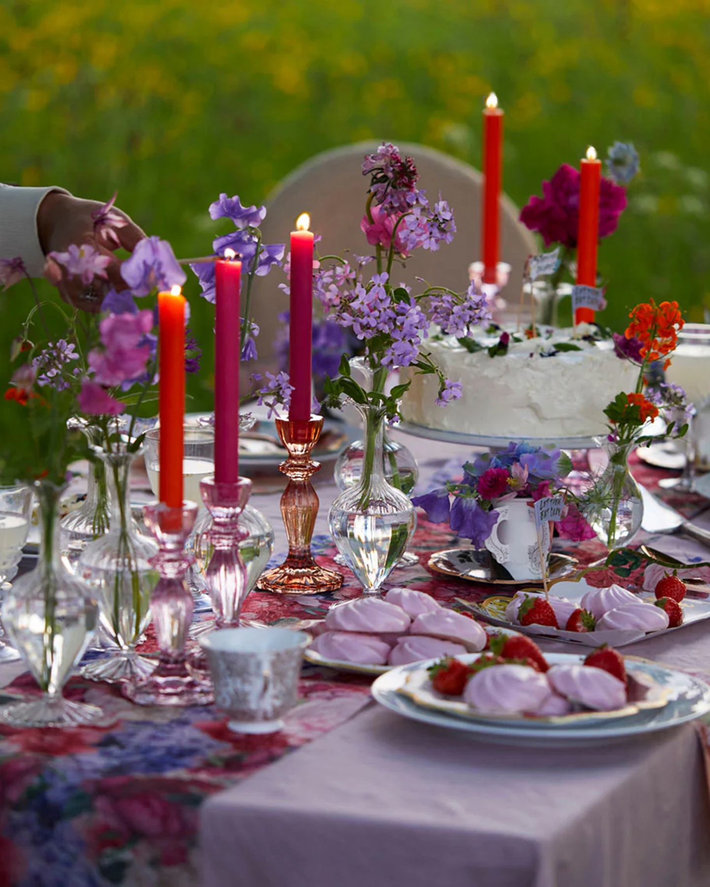 an outdoor table with a pink table cloth and a purple an dred floral runner, bright red lit dinner candles, bud vases with purple flowers and plates of strawberries and meringues