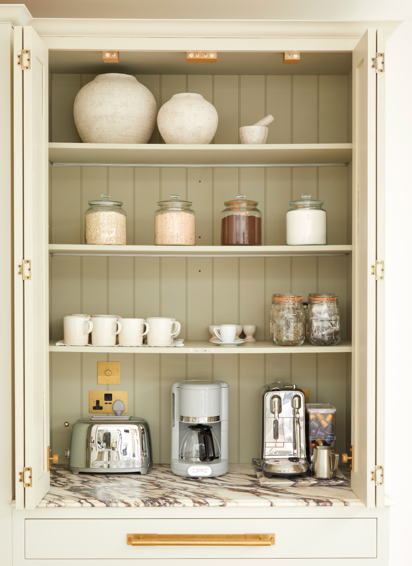 sage-green painted appliance larder with tongue-and-groove backing, open shelving displaying glass canisters and ceramics, brass sockets, and an espresso machine on a marble workto