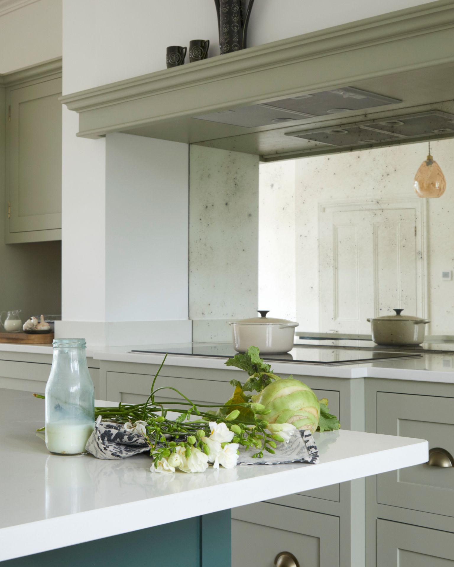 a kitchen with a large mirror splashback, sage green cabinetry and cooker hood and a white marble kitchen island top with milk and flowers on top