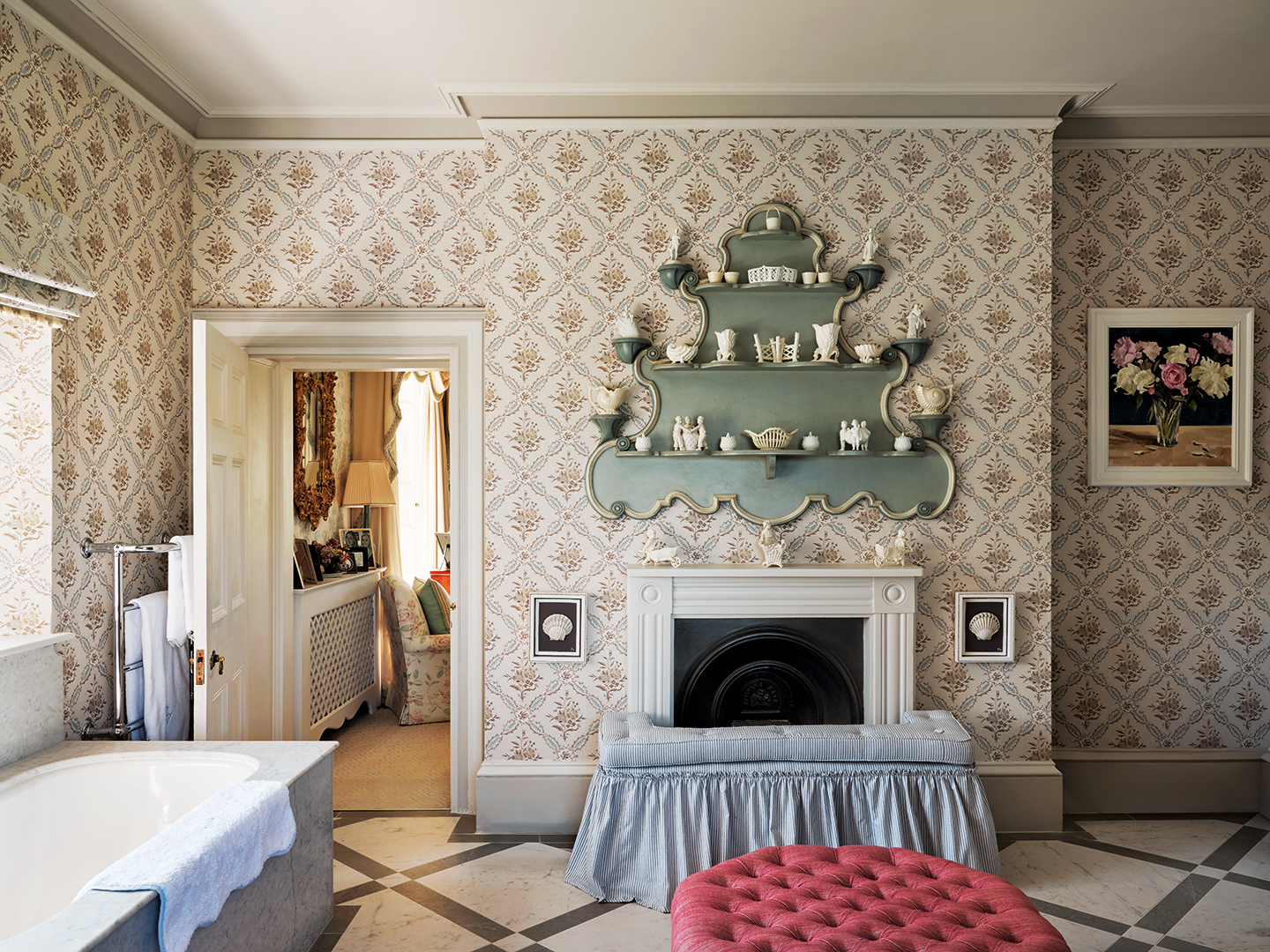 A large wallpapered bathroom with a central fireplace surrounded by an upholstered, skirted fender and above hangs an ornately-shaped set of shelves displaying various ceramics and objects. In the foreground is a red ottoman and a marble bath