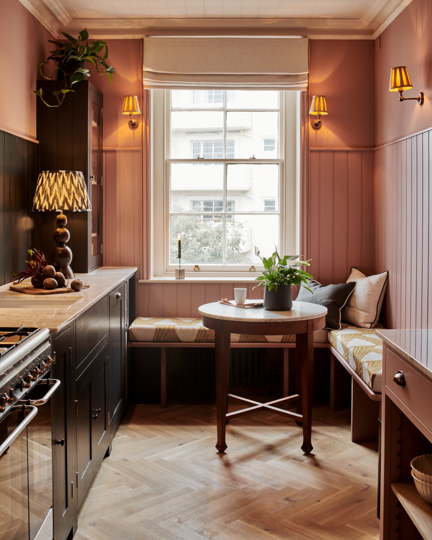 a kitchen with salmon pink walls, banquette seating under the window with a small round table, herringbone wooden flooring, black kitchen cabinets and pleated wall lights