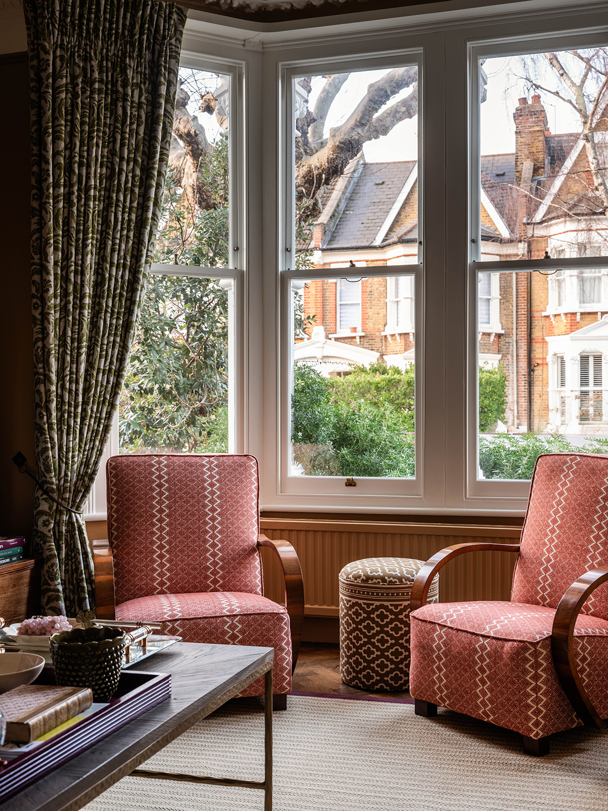 Two armchairs covered in rust red fabric with a diamond pattern sit in the bay window of a Victorian living room on a neutral rug, framed by tall curtains with a green damask floral pattern
