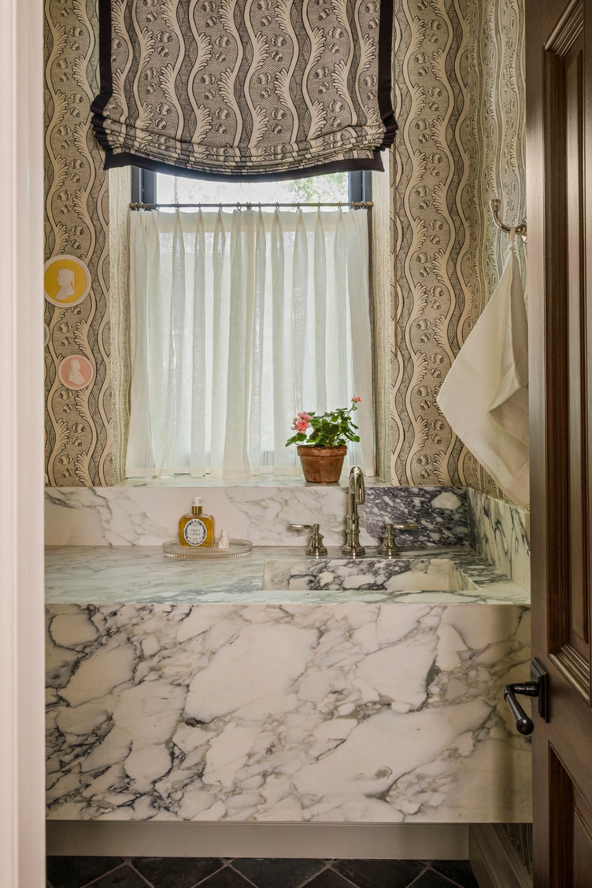 A small bathroom with a marble vanity unit, striped wavy wallpaper and matching relaxed blind, with a cafe curtain underneath. A flower pot and hand soap sit bedside the sink. Small circular intaglios hang on the wall 