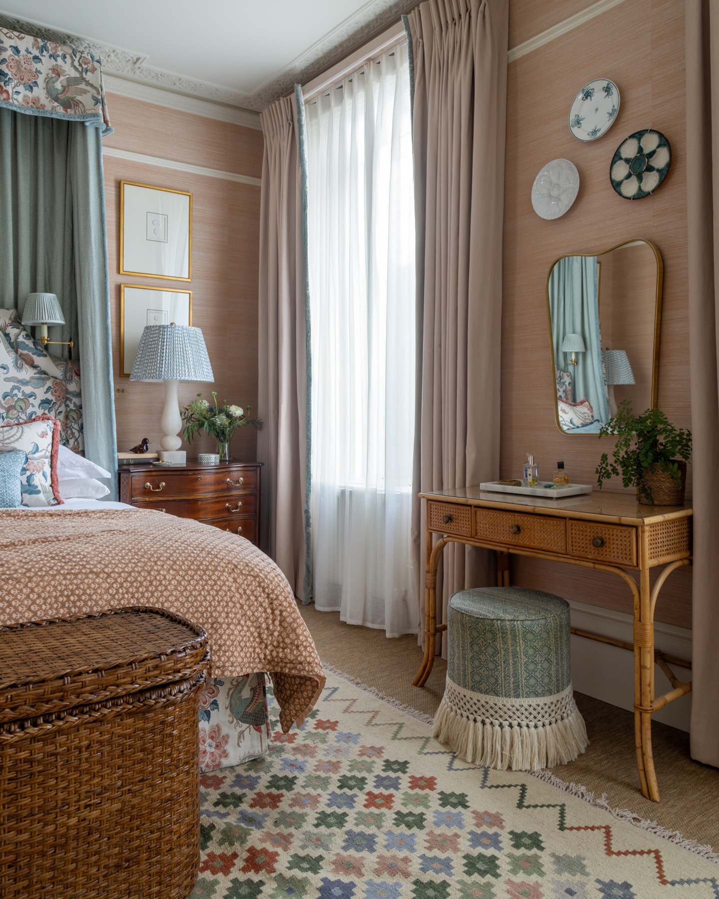 a bedroom with a rattan dressing table and ottoman, a vintage mirror above the dressing table, a vintage chest of draws next to the bed and a bed with a canopy and a beige and orange patterned bedspread