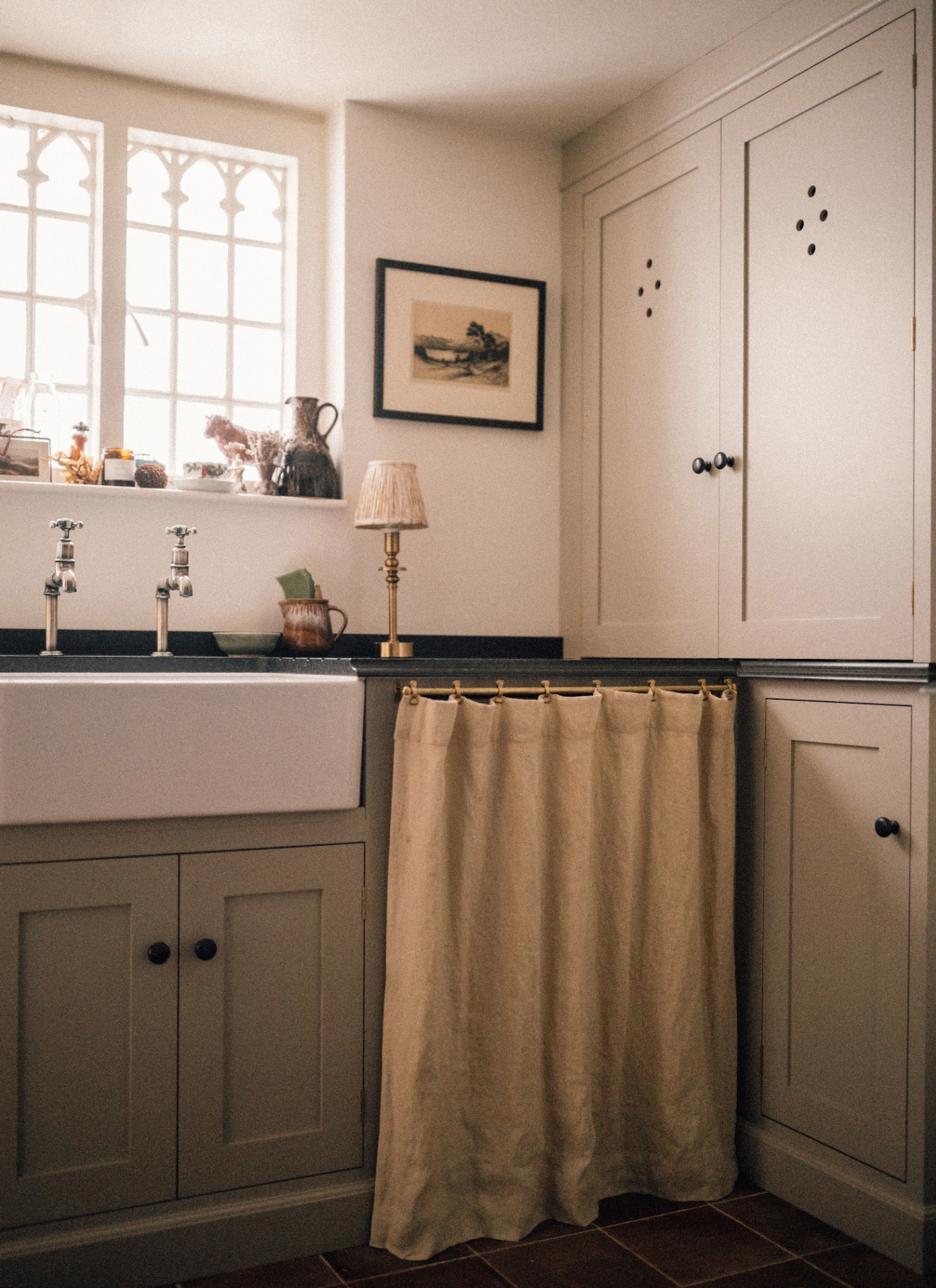 A kitchen corner with white walls and white cabinetry, a cream sink skirt, a small landscape painting on the wall and decorative objects on the window ledge