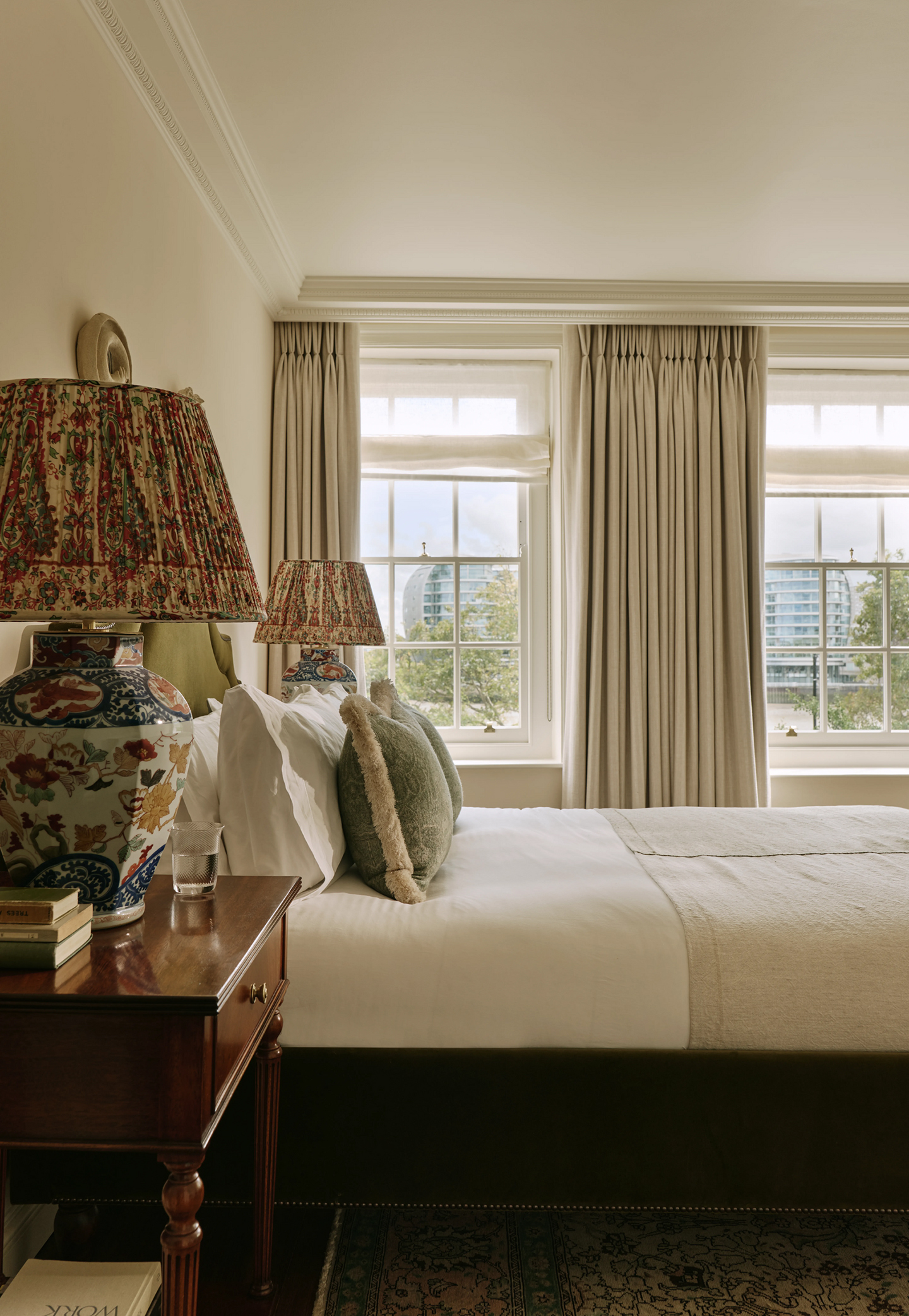 A side-on view of a bedroom with a double bed upholstered in green velvet fabric. A wooden bedside table sits in the foreground topped with a large chinoserie-style table lamp, books and a glass. Neutral curtains dress large sash windows on the back wall 
