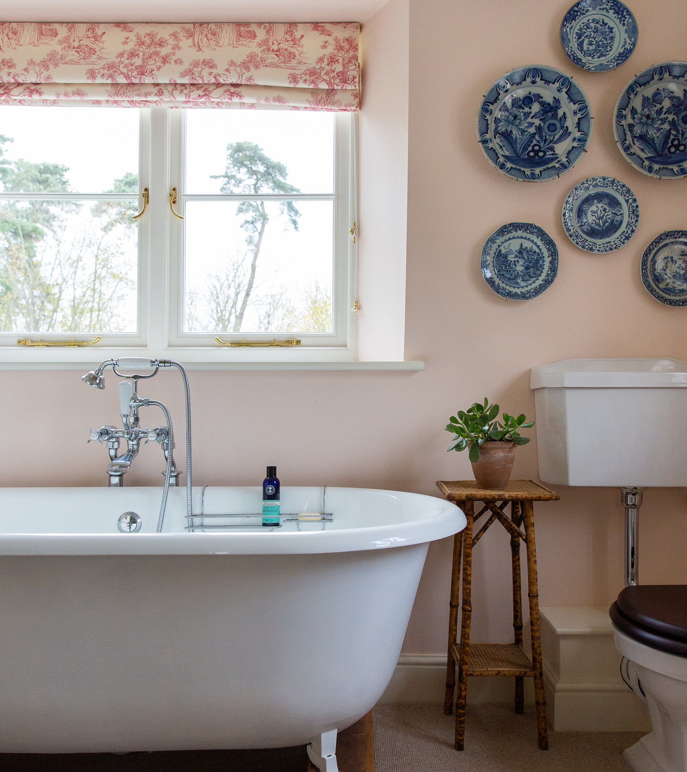 a bathroom with plaster pink walls and blue decoratie plates hung above the toilet, a freestanding bath infront of a large window with red and white patterend blinds