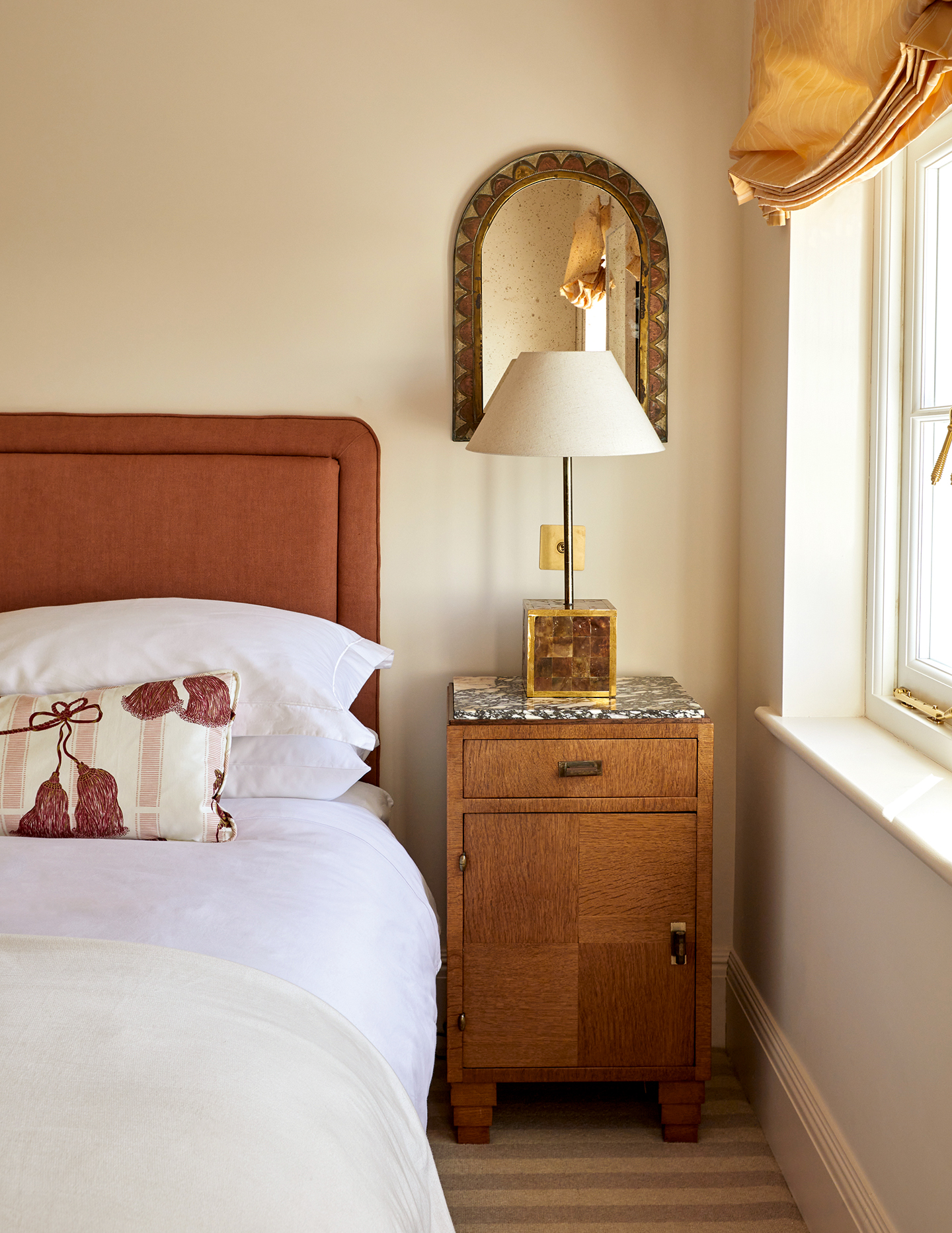 A bedroom with neutral walls, a brick red-toned upholstered headboard and a wood bedside table with a marble top holding a square table lamp. An arched decorative mirror hangs above