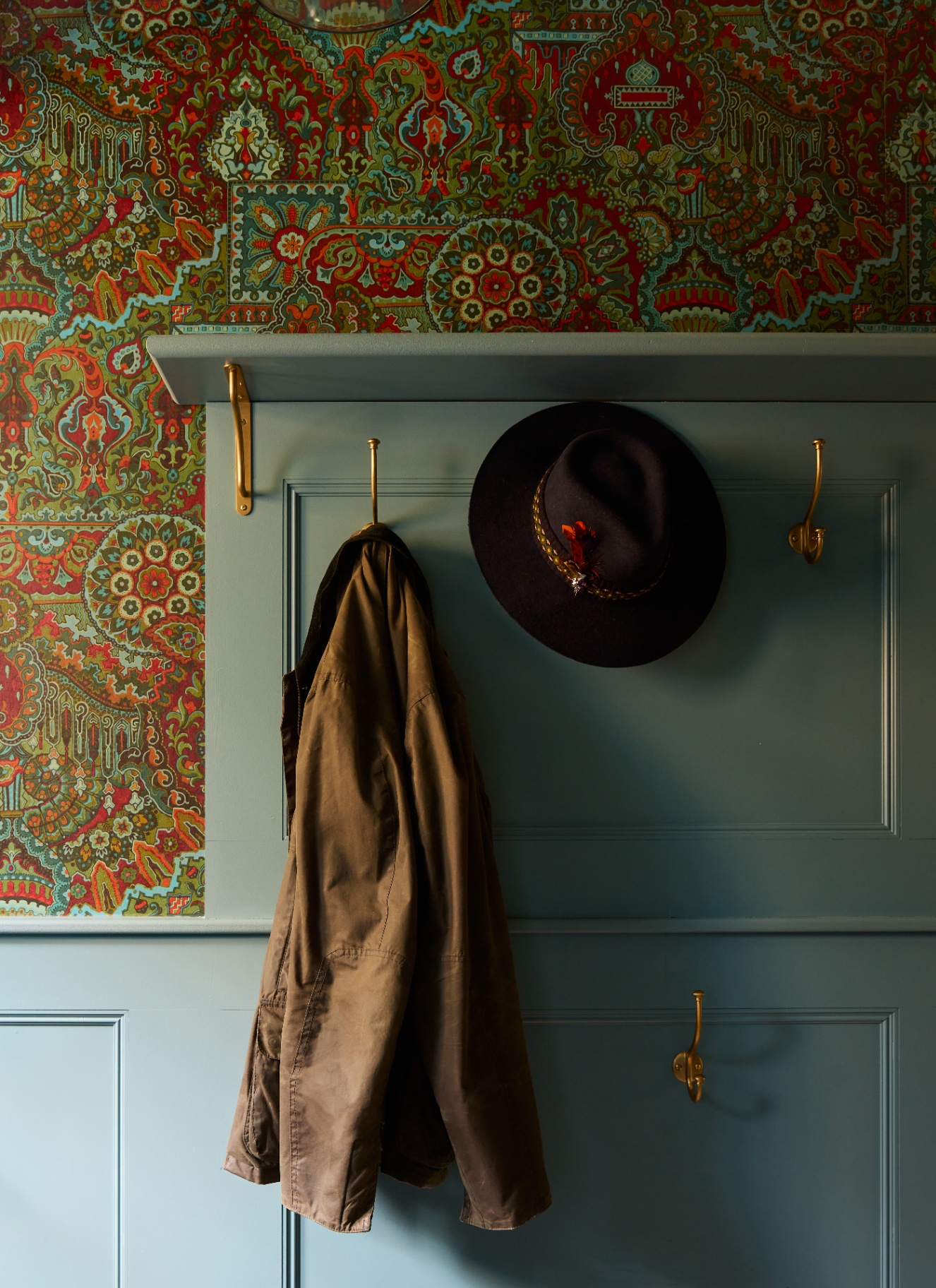 a close up of a hallway with blue painted walls with gold coak hooks with a coat and a hat hanging and a bright patterned wallpaper