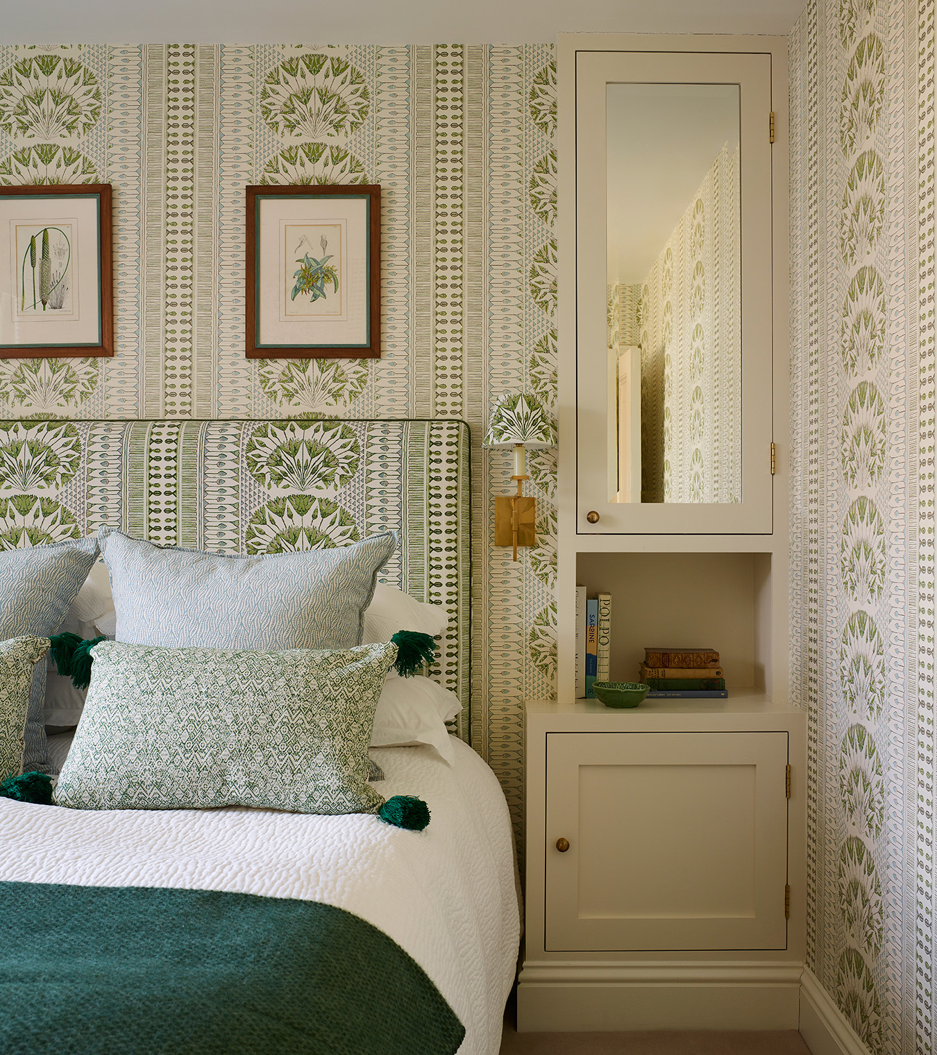 A bedroom with a double bed with white bedding and green patterned cushions, a built in cupboard with a mirror, white and green botanical wallpaper on the walls and a matching patterned headboard