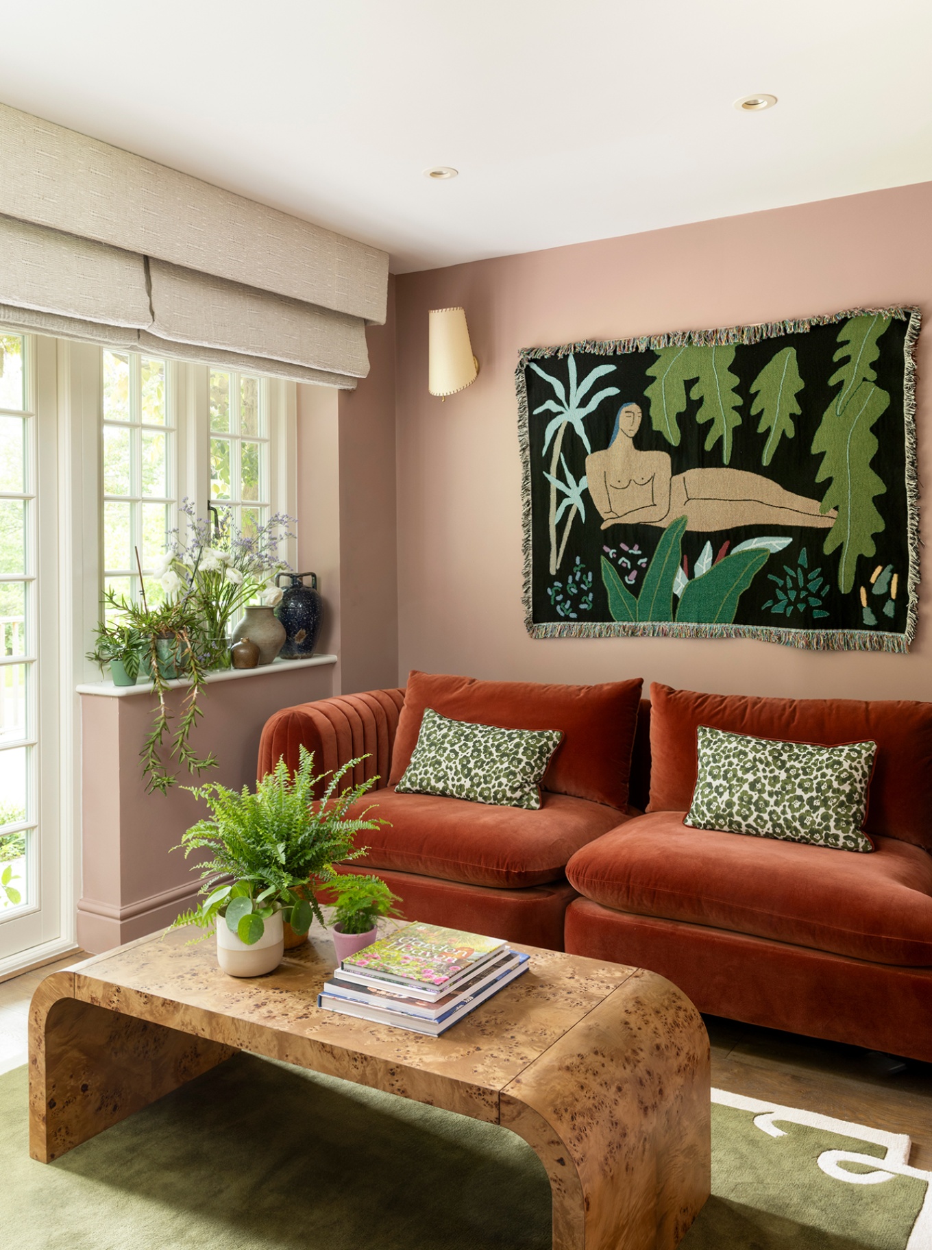 a garden room with pink walls, orange velvet sofa and arched burl wood coffee table set on a green rug. Doors and windows leading to the garden line the left wall and on the back wall hangs a conical wall light and botanical wall hanging