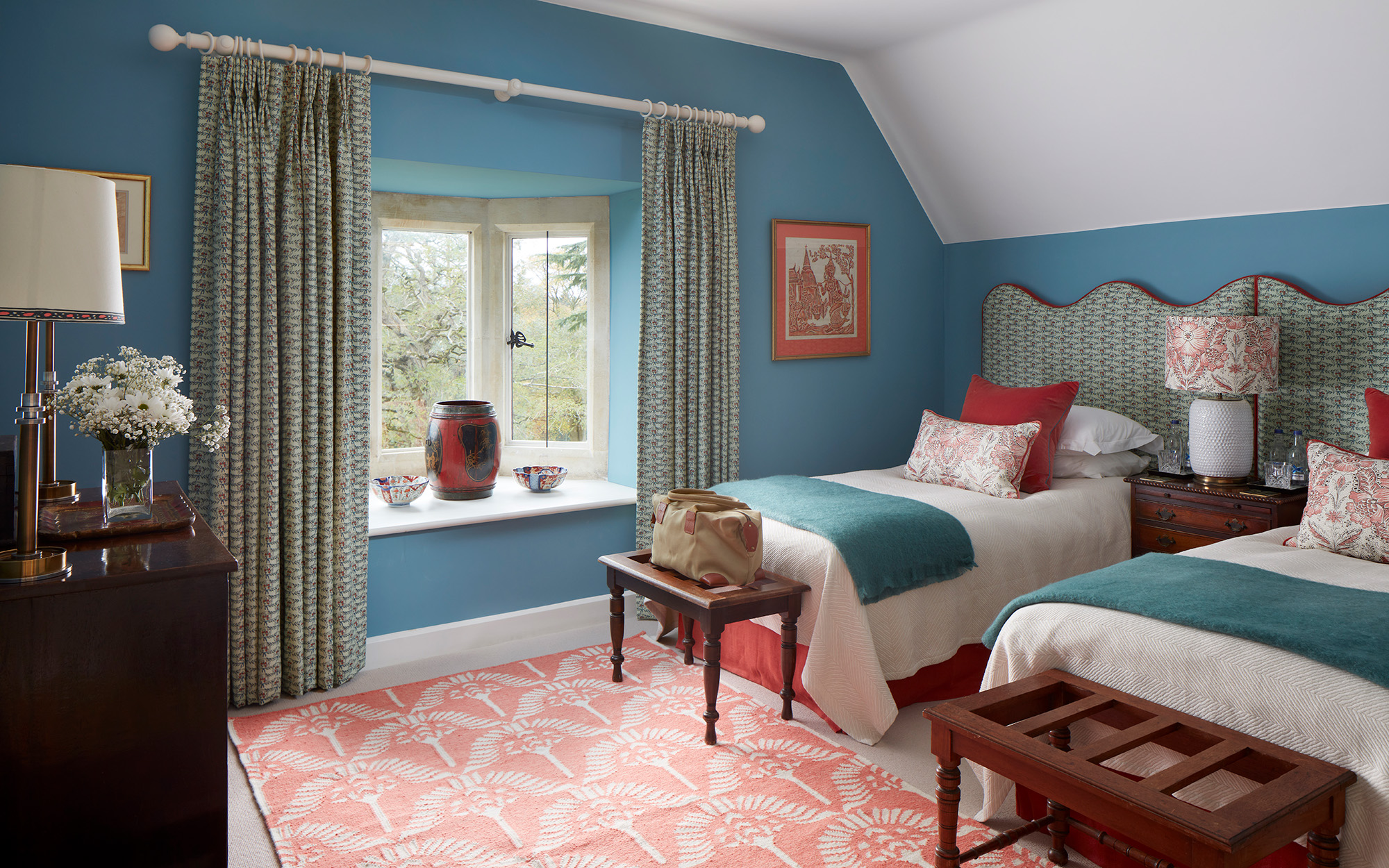 a blue bedroom with twin beds, each with a green patterned headboard, white bedding, a red and a floral cushion and a teal throw, a wooden bench at the end of beds, a coral rug, a mahogany chest of drawers and a window with green curtains