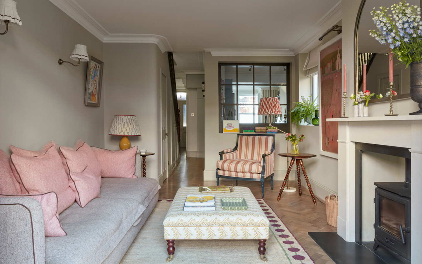 A light grey sofa with soft pink pillows sits across from a black wood-burning stove in a neutral living space. A patterned ottoman on a border-trimmed rug, a striped armchair, and a tripod side table complete the room.