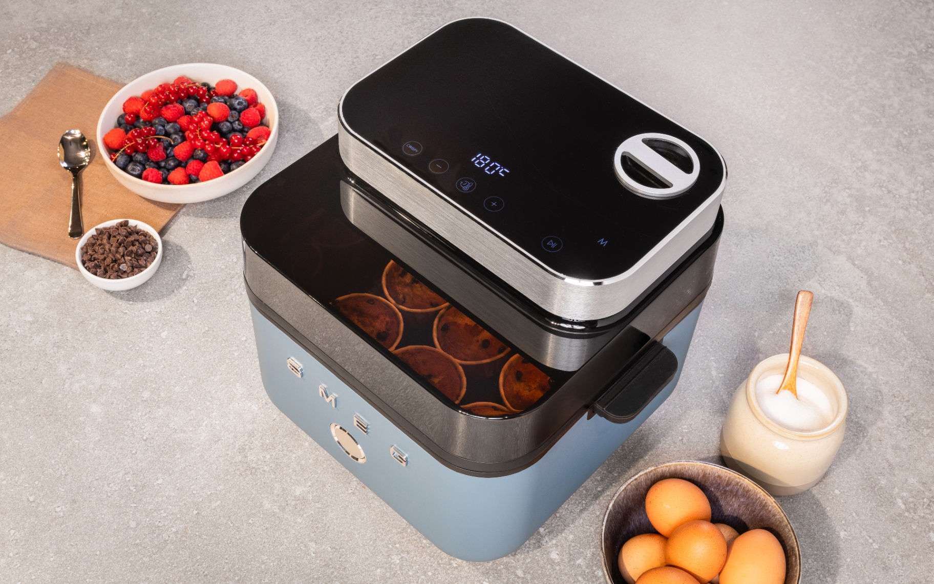 High-angle view of a pastel blue SMEG air fryer baking small cakes visible through its glass window. The top digital display shows 180°C. Surroundings include bowls of fresh berries, chocolate chips, eggs, and sugar on a grey countertop