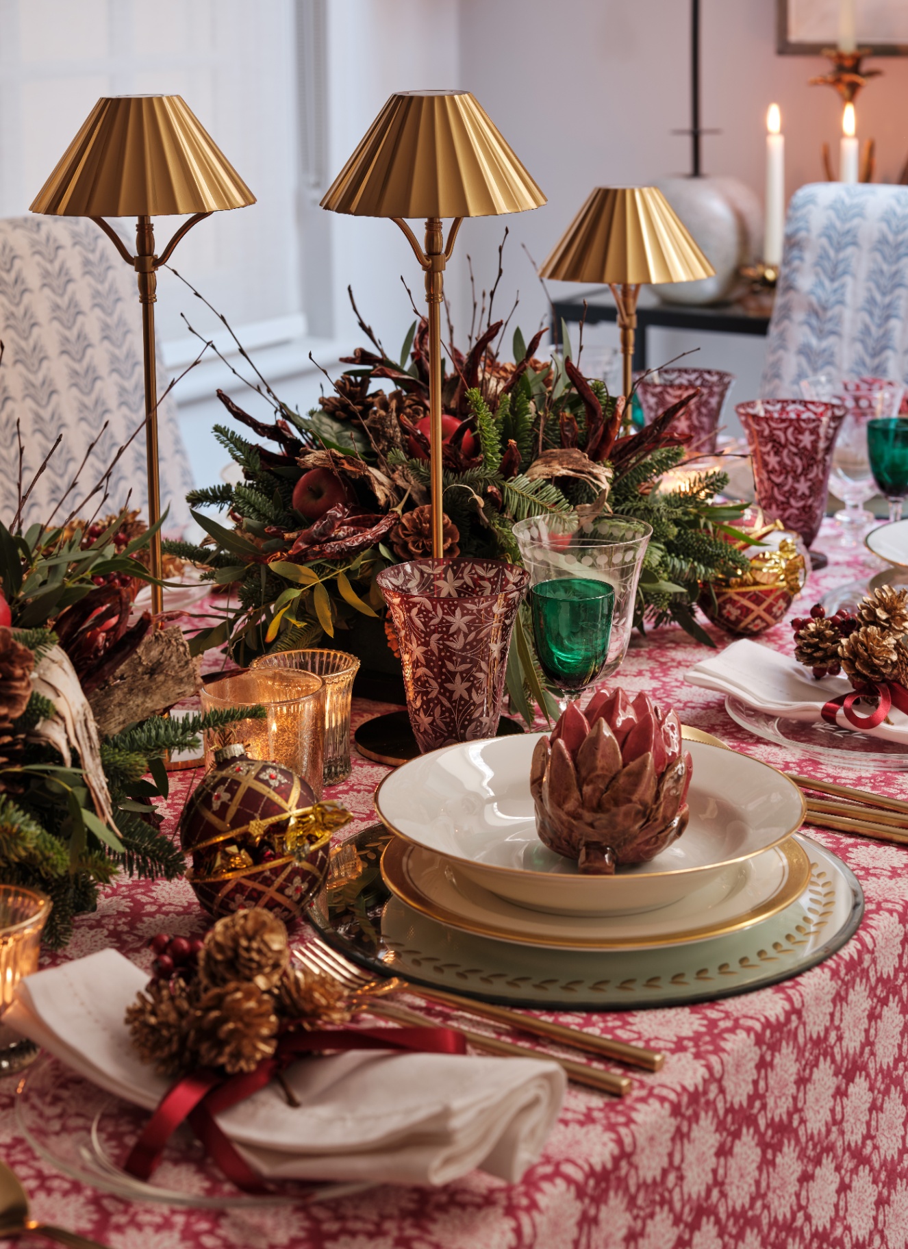 A festive dining table with a red patterned tablecloth, brass mini lamps, glassware, and a centerpiece of dense spruce, pinecones, dried chilies, and holiday foliage