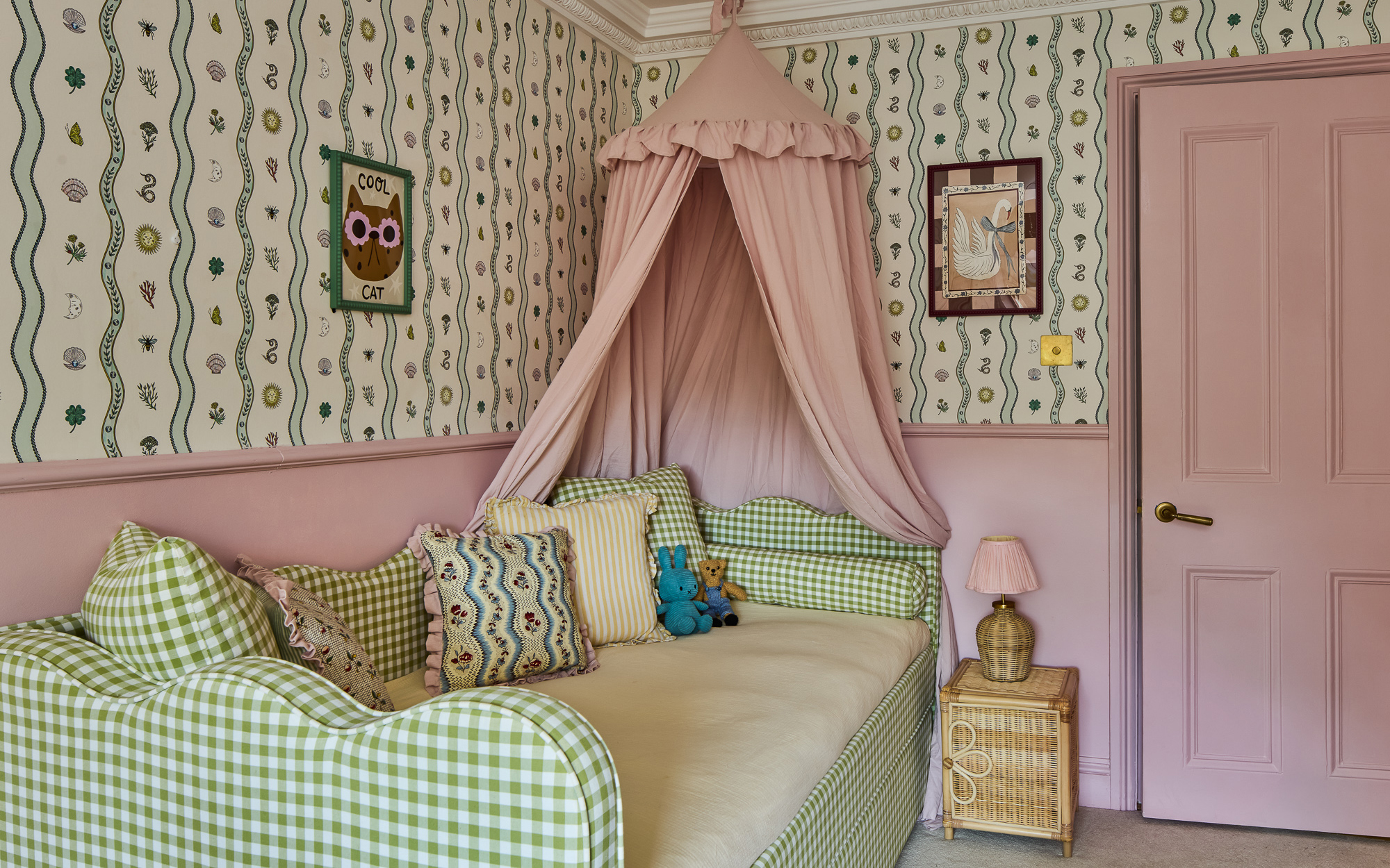 A cozy bedroom with a green-and-white gingham daybed under a dusty-pink canopy. The room features matching pink lower walls and door, vintage-style patterned wallpaper, framed art, and a small woven side table