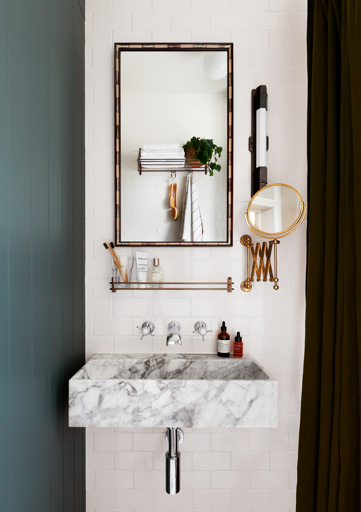 a close up of a wall-mounted marble basin paired with wall-mounted silver taps, on a white tiled bathroom wall. Above hangs a striped mirror, brass mounted shelf, an extendable shaving mirror and a narrow wall light