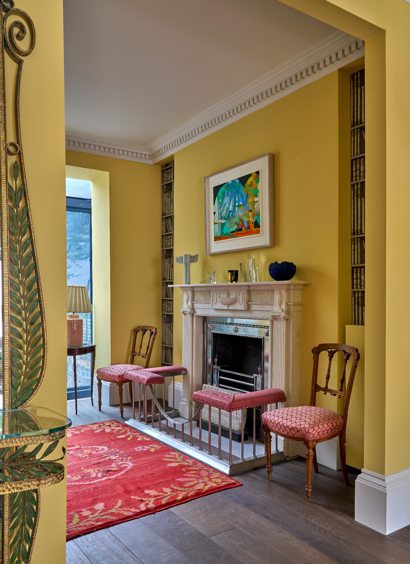 a sitting room with bright yellow walls and white ceiling and cornicing, a large white fireplace and two wood and red dining chairs next to it