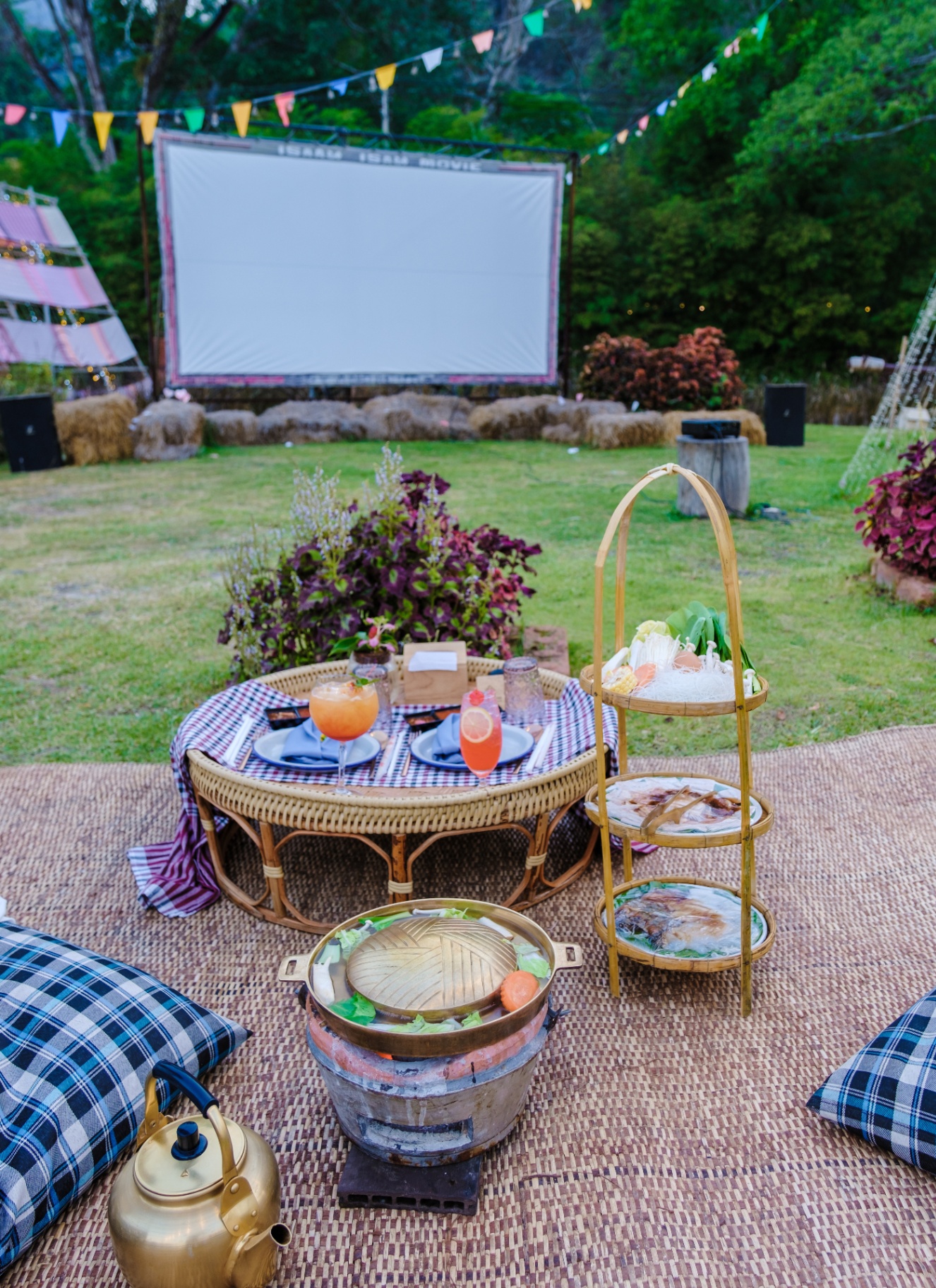 an outdoor cinema screen on a lawn with a jute picnic rug with cushions, a table and drinks on it
