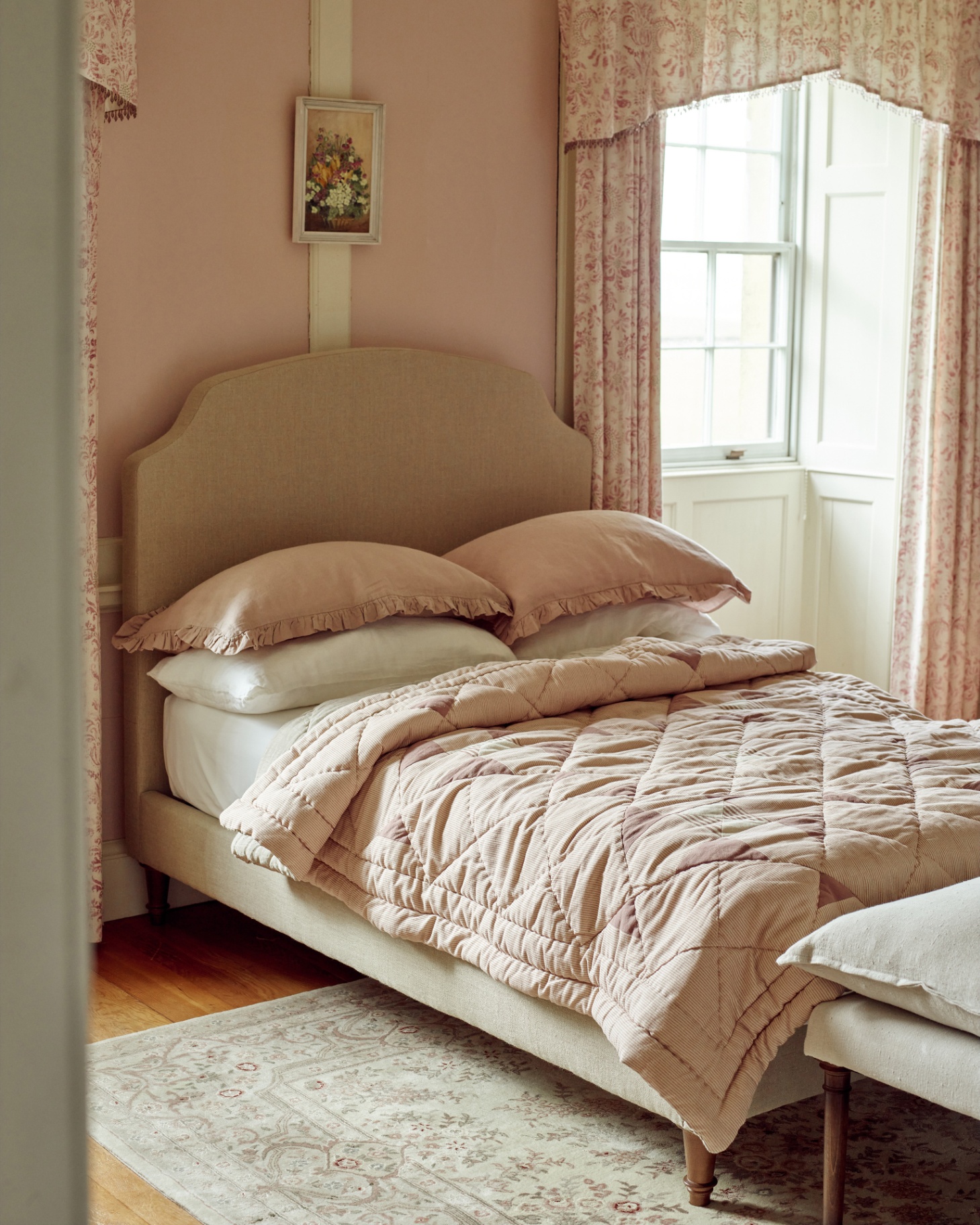 a bedroom with a double bed with pale pink bedding, pale pink walls and a large window with pink floral curtains