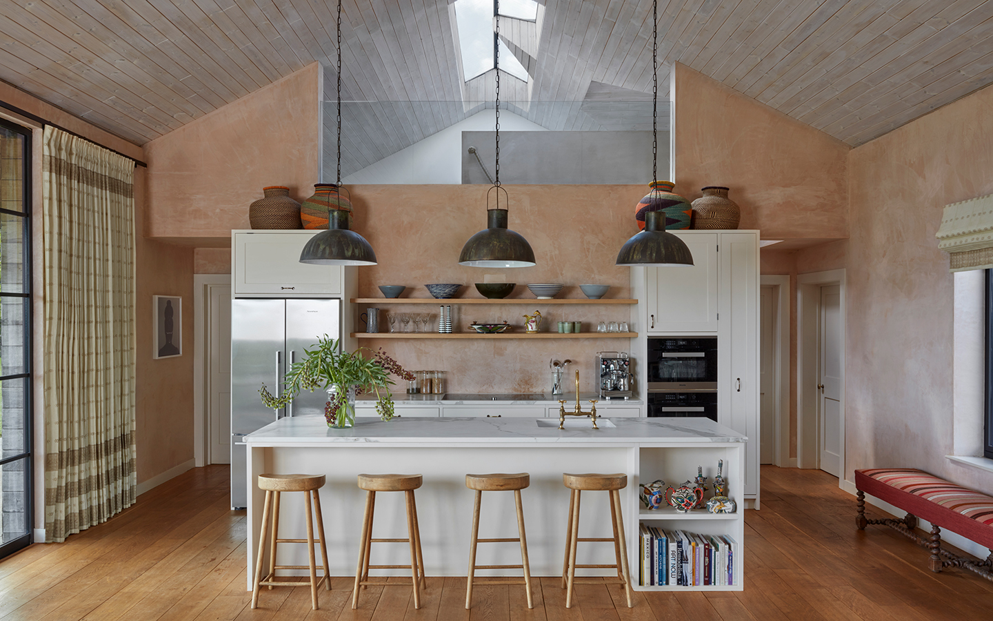 An airy kitchen with white, marble-topped island, paired with four wooden bar stools, and white cabinetry. Open wooden shelves hold kitchen utensils and ceramics. The tall ceilings are timber-clad and the walls are raw plaster. A mezzanine is seen above