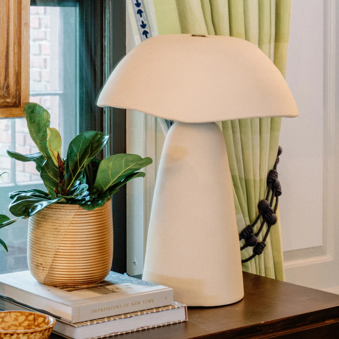 a cream stone effect table lamp on a wooden table next to a pile of books and a plant in a gold ribbed pot in front of pale green curtains