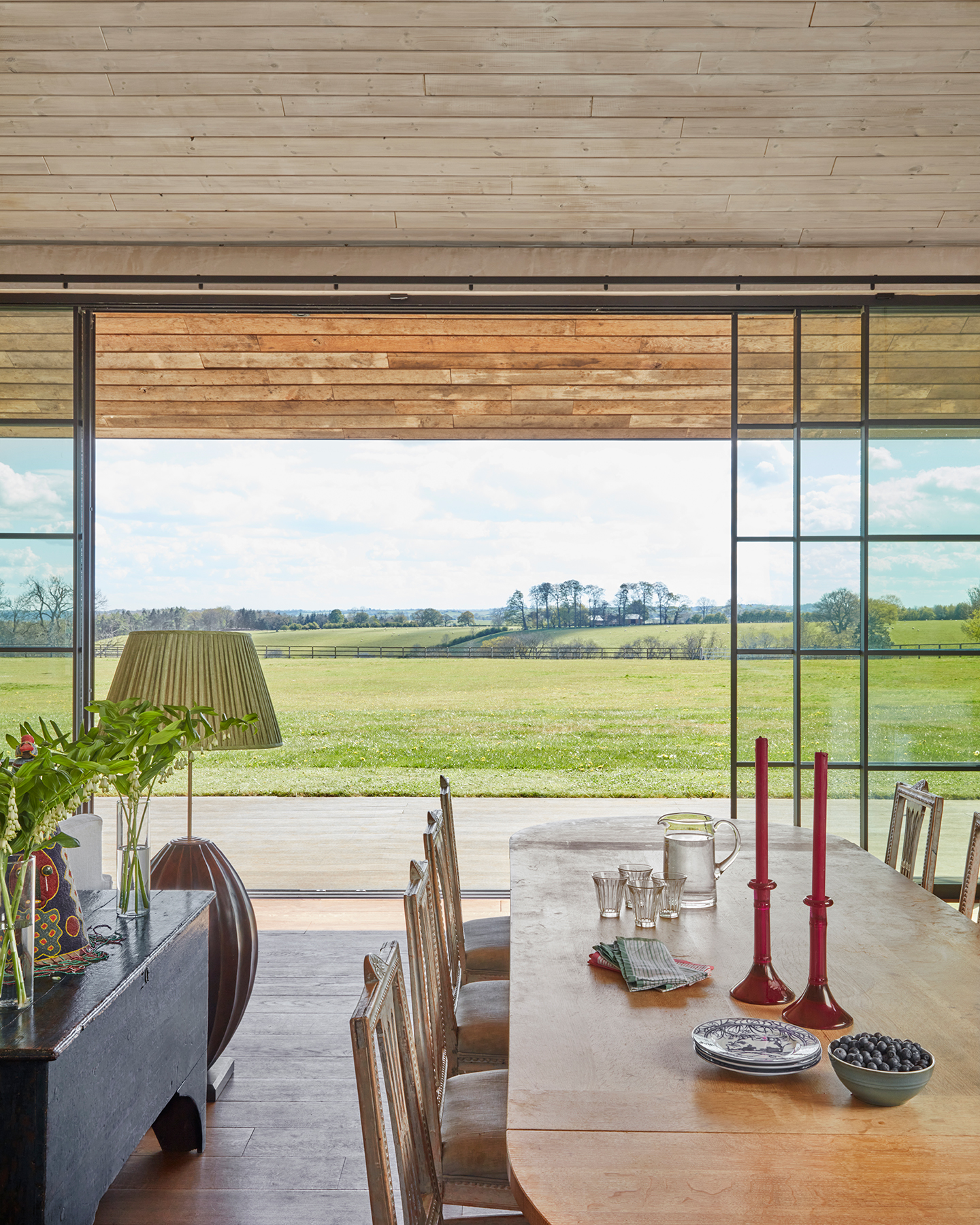 In the foreground, a wood dining table is surrounded by chairs and topped with ceramics, glassware and candlesticks. The back wall is glazing and french windows are open. Rolling hills can be seen beyond the garden