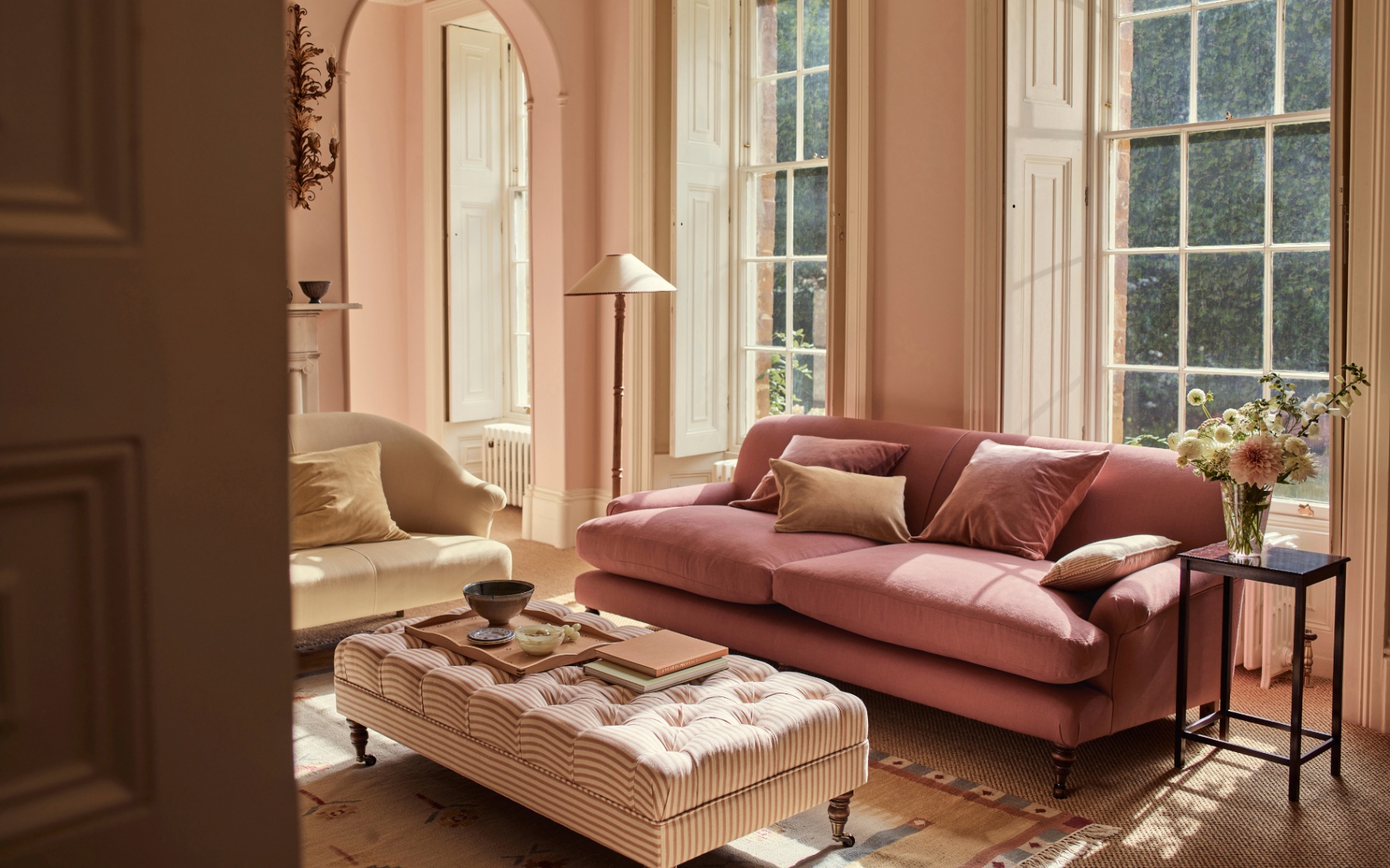 a living room with a pink velvet sofa in front of large windows and a cream and red stripe ottoman