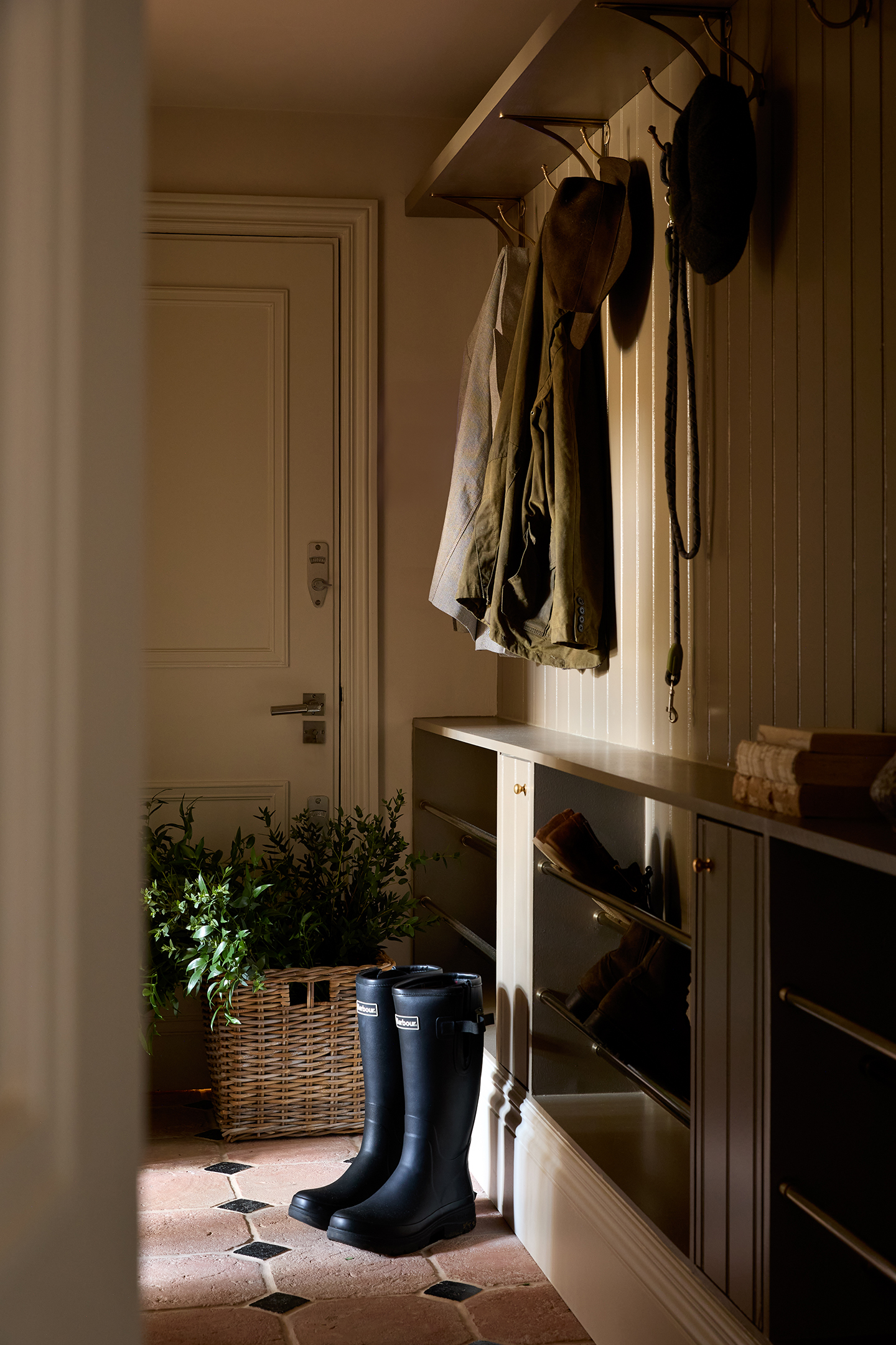 A boot room with tongue and groove panelling, an open shelf with built-in coat hooks and bespoke shoe storage on one wall. Stone tiles with a diamond pattern cover the floor. A pair of wellington boots and basket of foliage sit on the floor