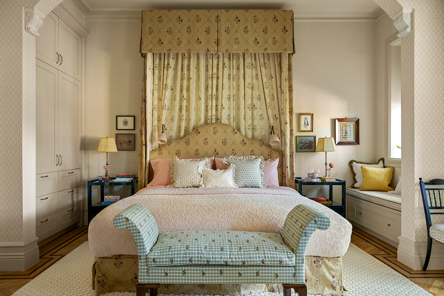 A bedroom with high ceilings. A large double bed is topped with a tall half-tester. At the end of the bed, is a floral check bench with scroll arms. A built-in bench sits to the right and and a built-in wardrobe to the left