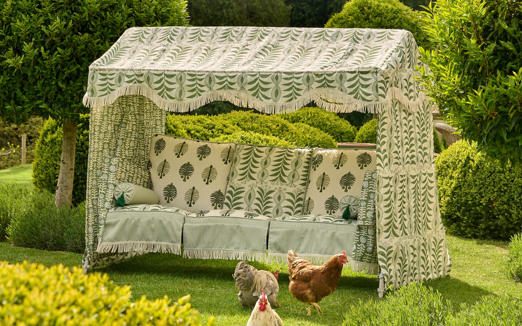 an outdoor bench covered with a patterned canopy and green and white patterned cushions, surrounded by trees and greenery and chickens in the foreground