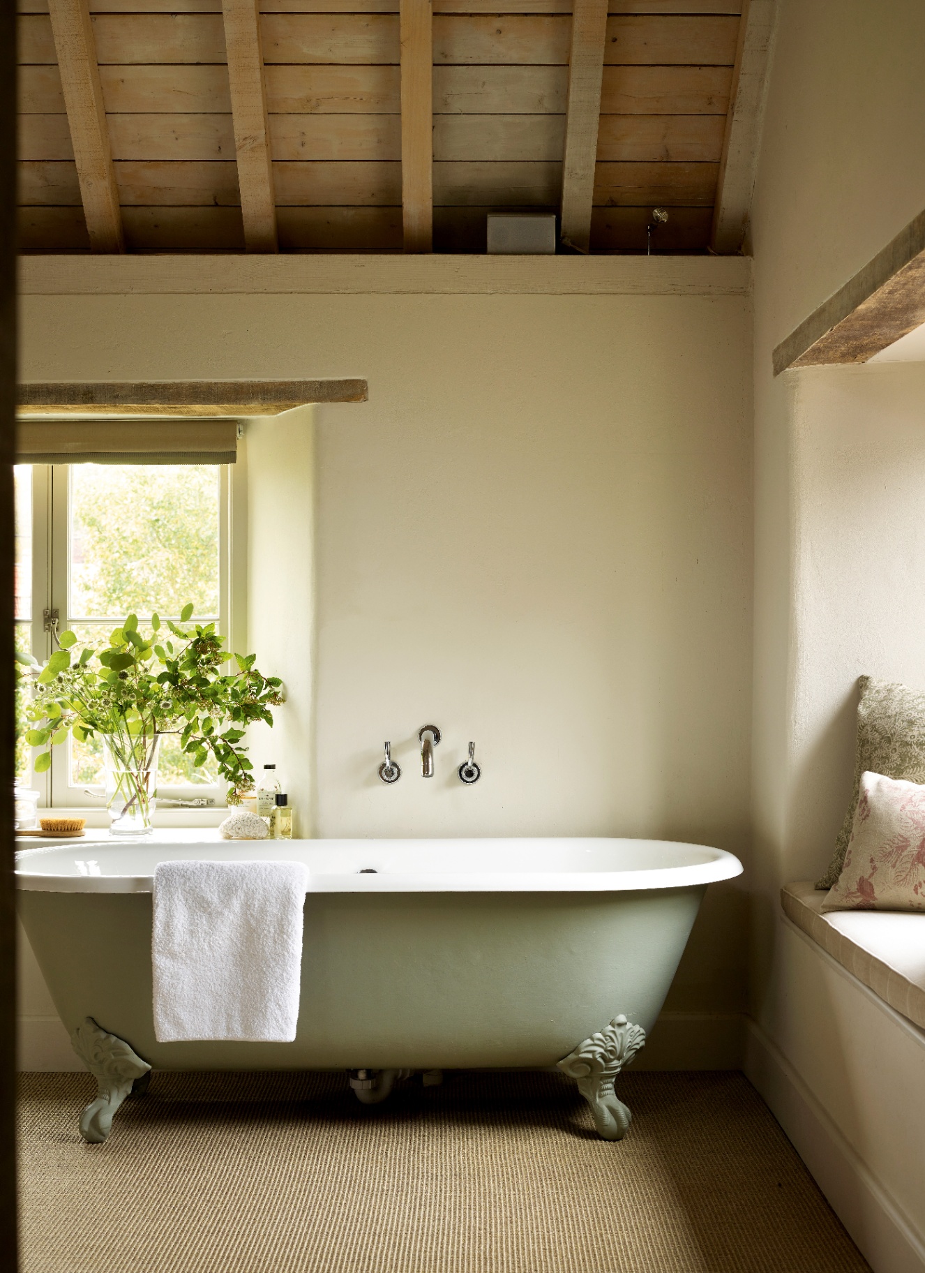 a neutral bathroom with a pale green freestanding bath, natural carpet, exposed beams and a vase of green leaves on the window ledge