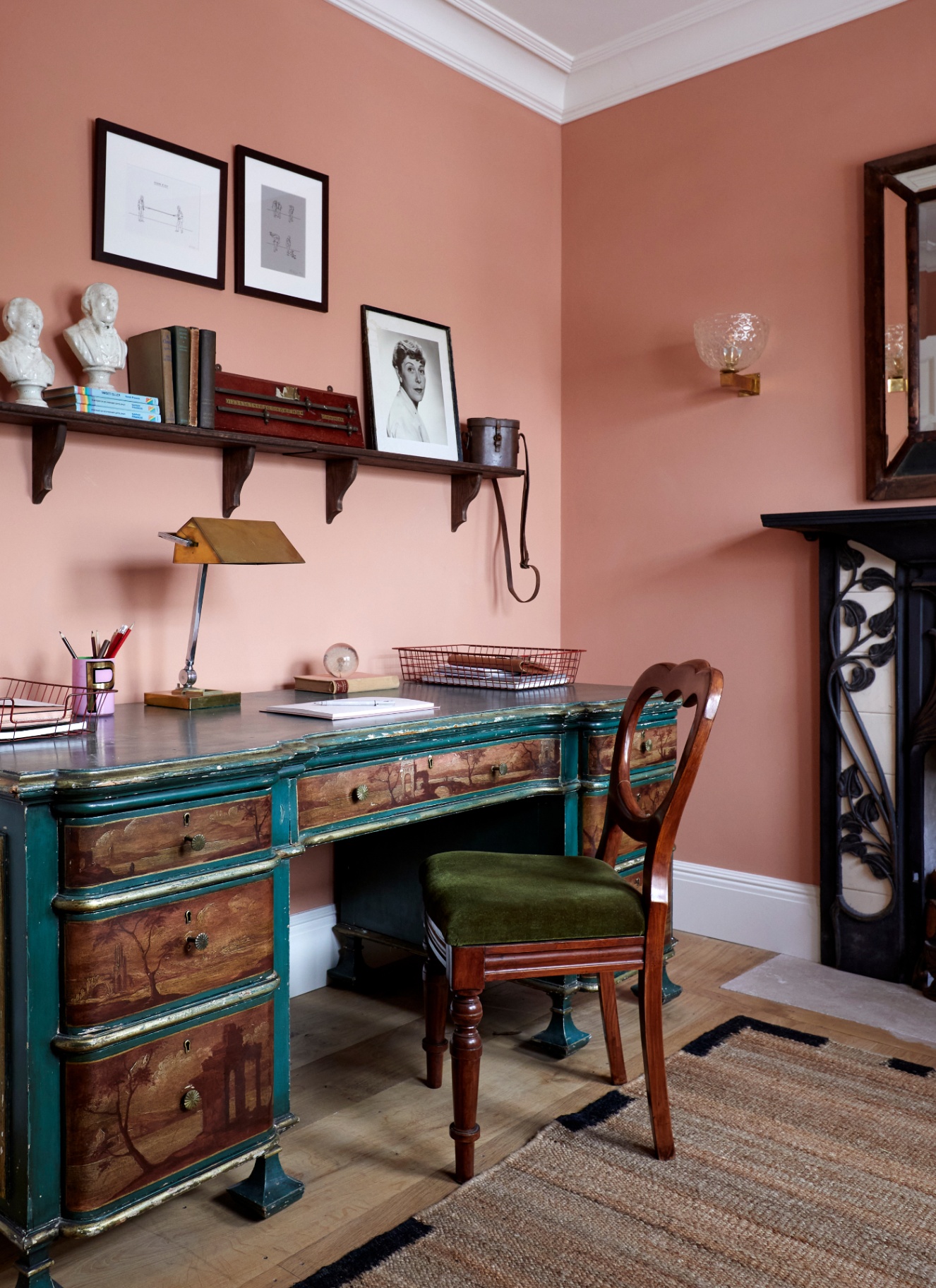 Warm peach-pink home office with a dark wood desk, green velvet upholstered dining-style desk chair, wall shelf, brass lamp, and cast-iron fireplace