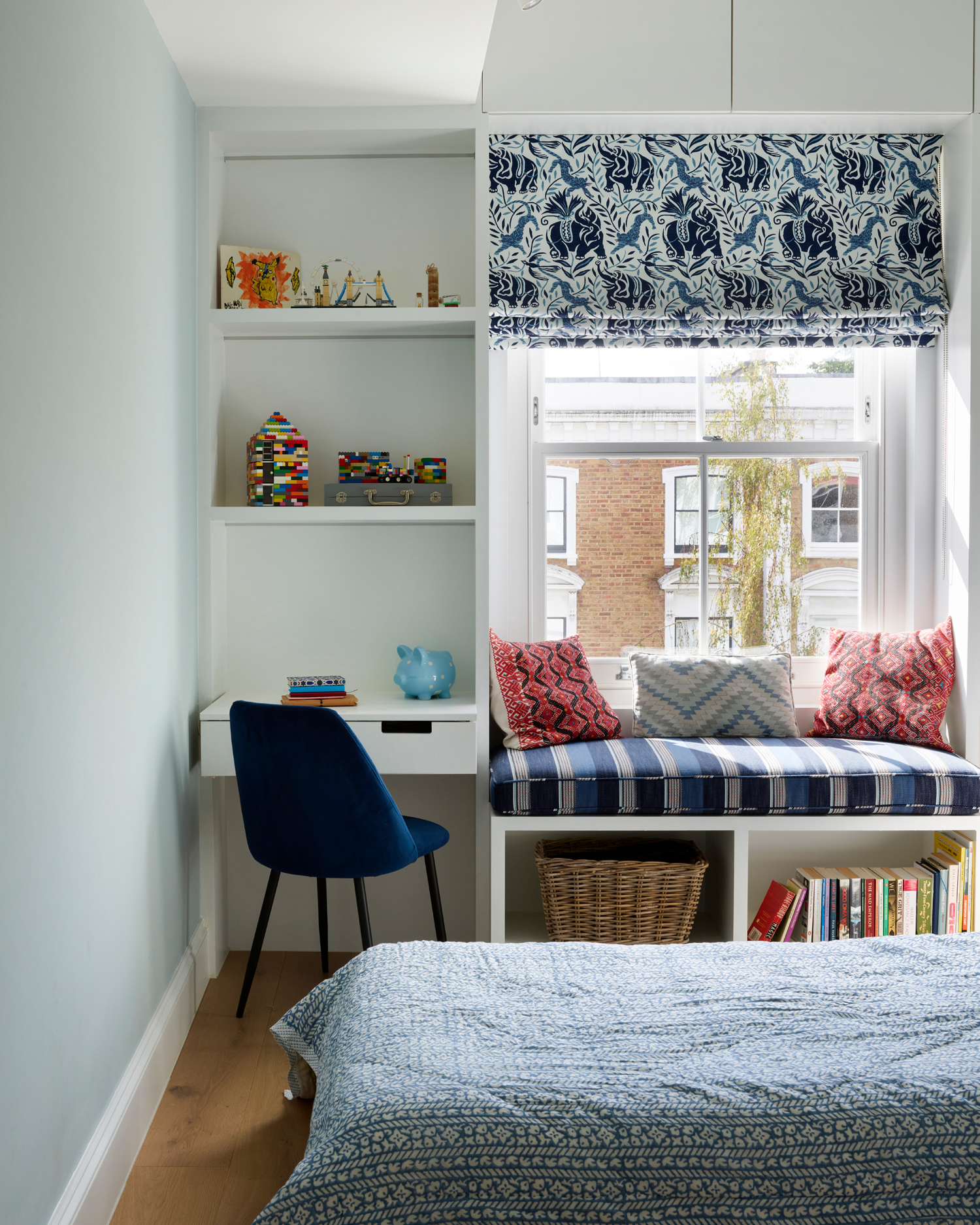 a bedroom with a built in desk and shelving unit, a window with an upholstered window seat with cushions on top and baskets and books underneath, and the end of a bed with a blue patterned bedspread