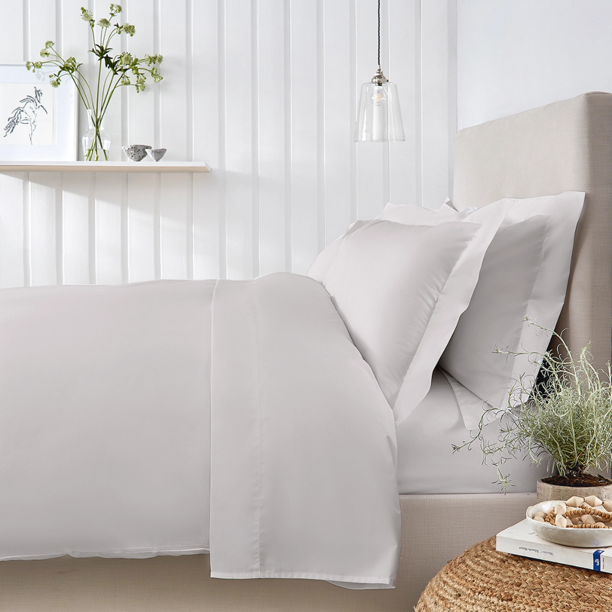 crisp white bedding on a double bed, a white paneled wall with an open shelf and a vase on and a jute footstool next to the bed with a plant and a book on top