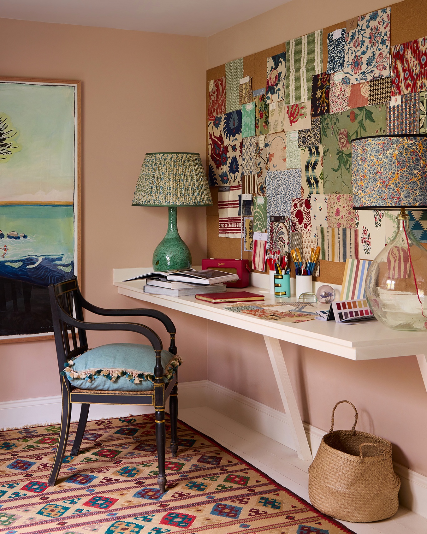A chair and desk in a pale pink home office. On the wall is a cork display board covered in fabric and wallpaper samples. 