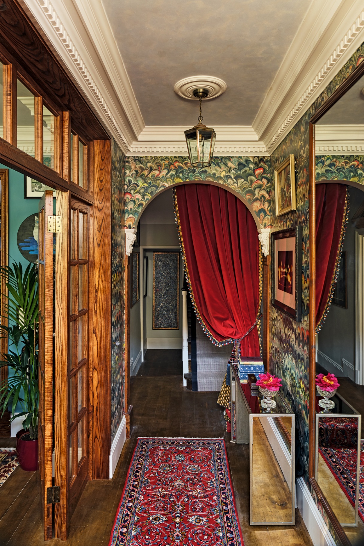 a hallway with marble wallpaper and an arched doorway towards the back dressed with a draped red velvet curtain. On the left are a set of glass-paned and wood internal doors and on the right a mirror and mirrored console table