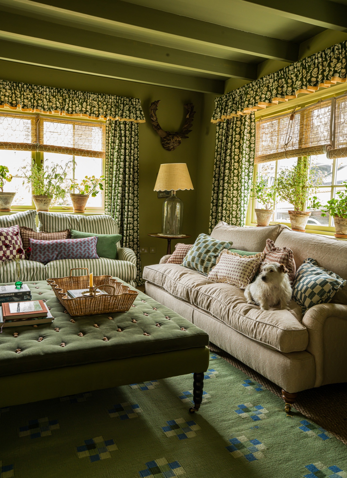 A richly decorated living room featuring an ochre velvet sofa, a large teal and red fabric ottoman, and a textured natural rug on herringbone wood floors. Vintage furniture, pattern-rich textiles, and a long framed art piece add depth