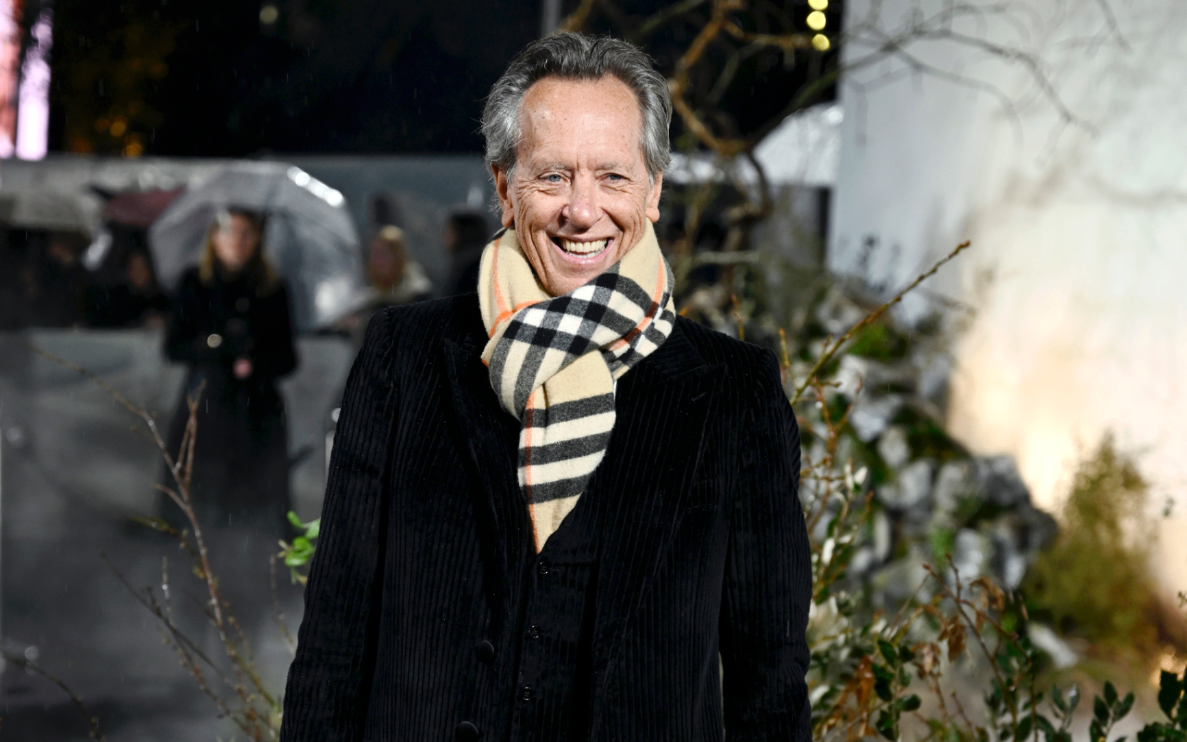 Actor Richard E. Grant smiling at an outdoor evening event, dressed in a black velvet or corduroy coat with a beige, black, and red Burberry plaid scarf wrapped around his neck against a blurred backdrop
