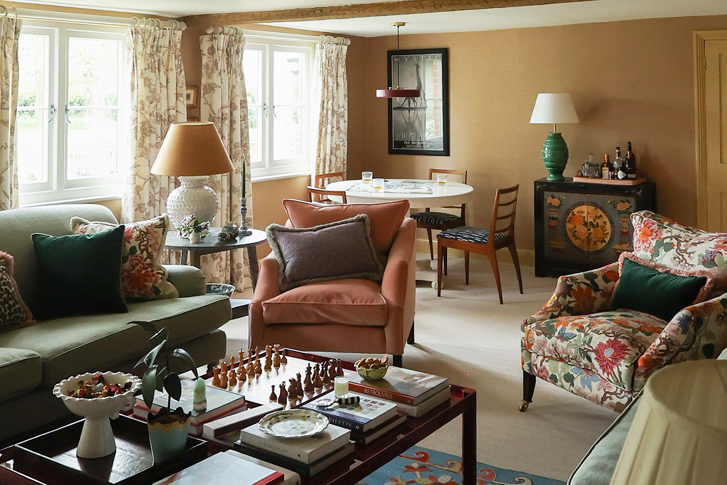 A sitting room with a seating area in the foreground, consisting of green sofas, a pink armchair and a floral armchair around a styled coffee table, and a dedicated games table with wooden dining chairs in the background