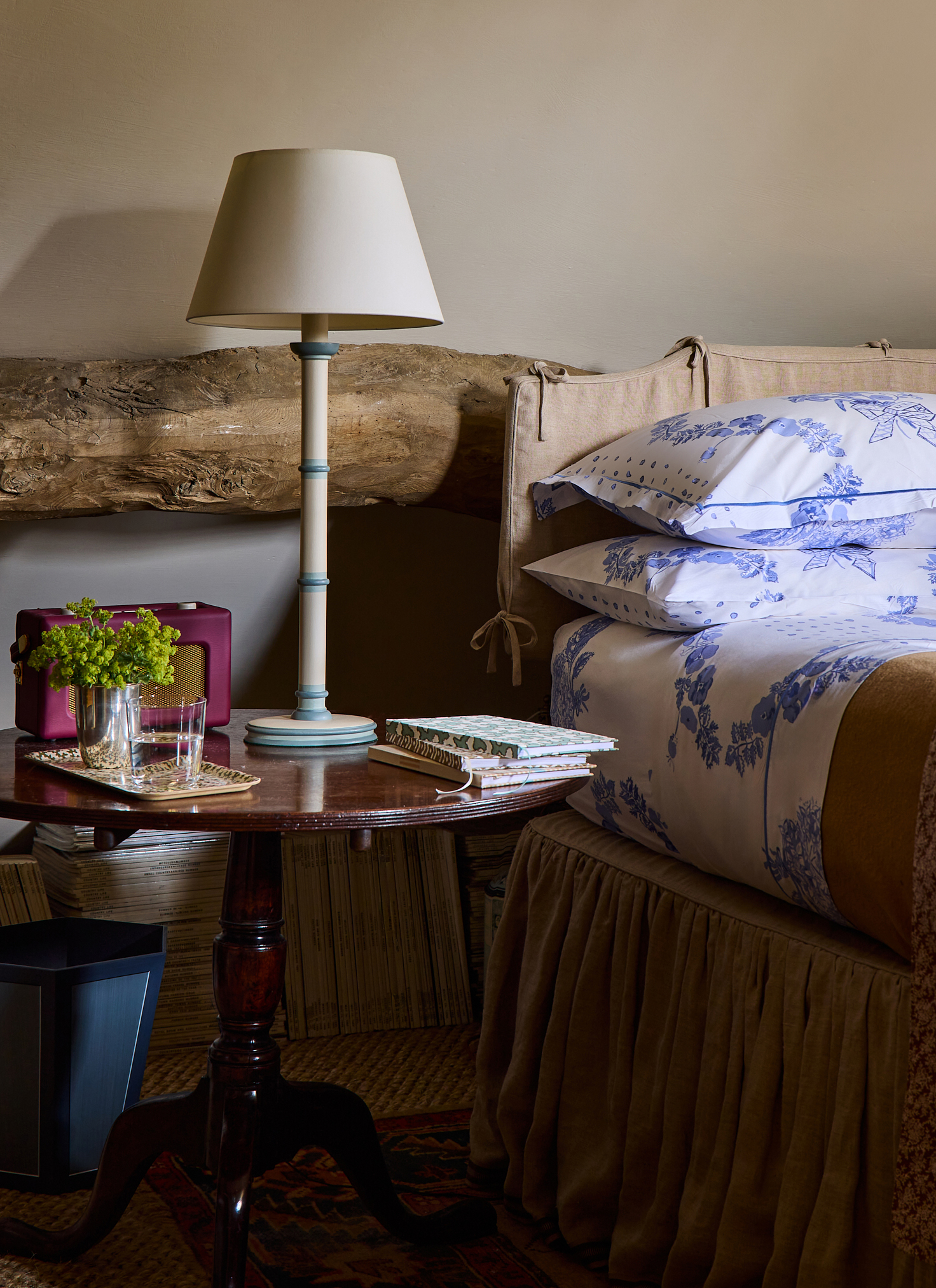 a bed with white and blue floral bedding and a wooden bedside table with a cream lamp and a red roberts radio