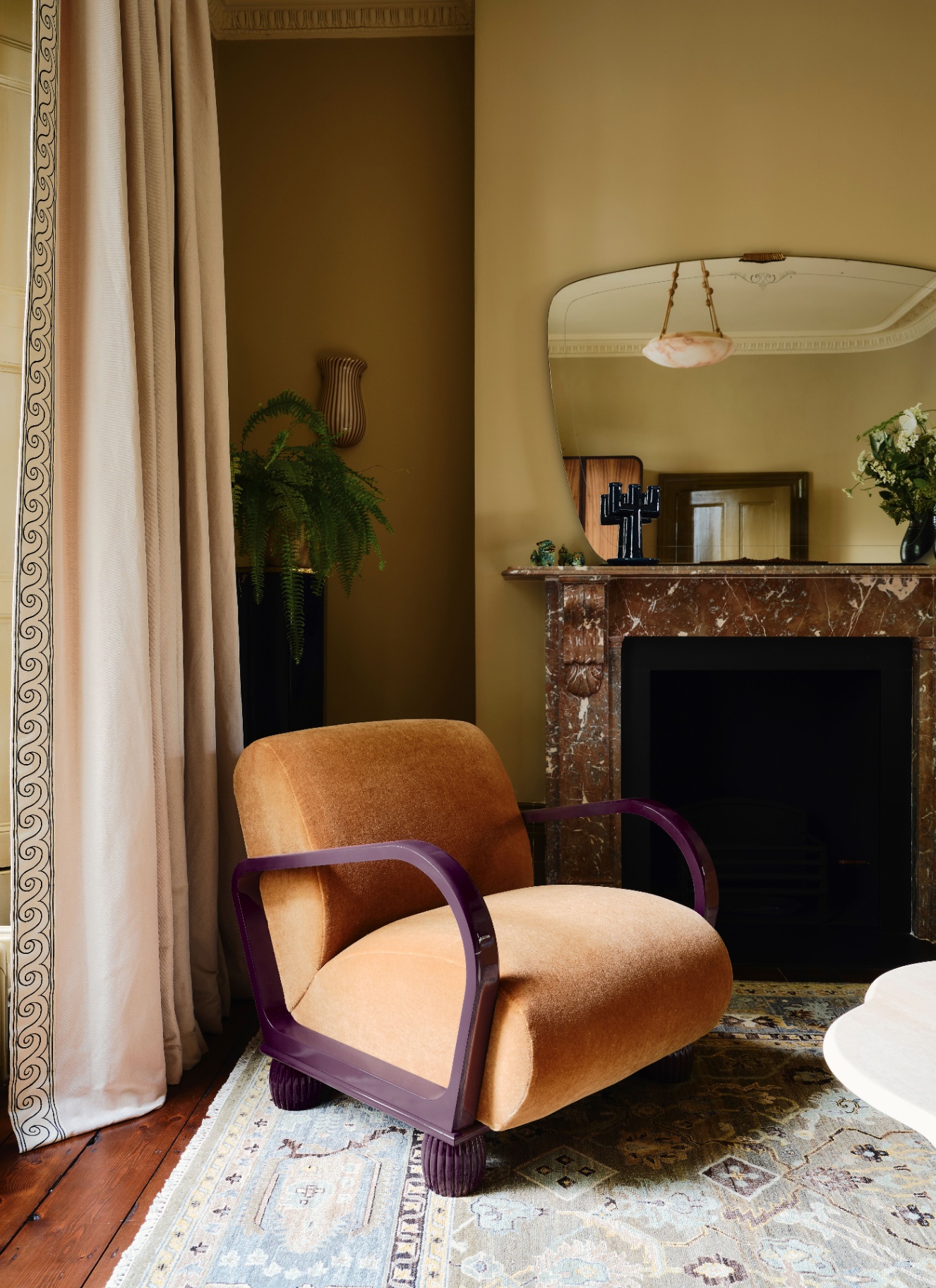 a sitting room corner with a statement chair in ochre with plum arms and legs, pale green walls, a large brown marble fireplace and an art deco mirror above it