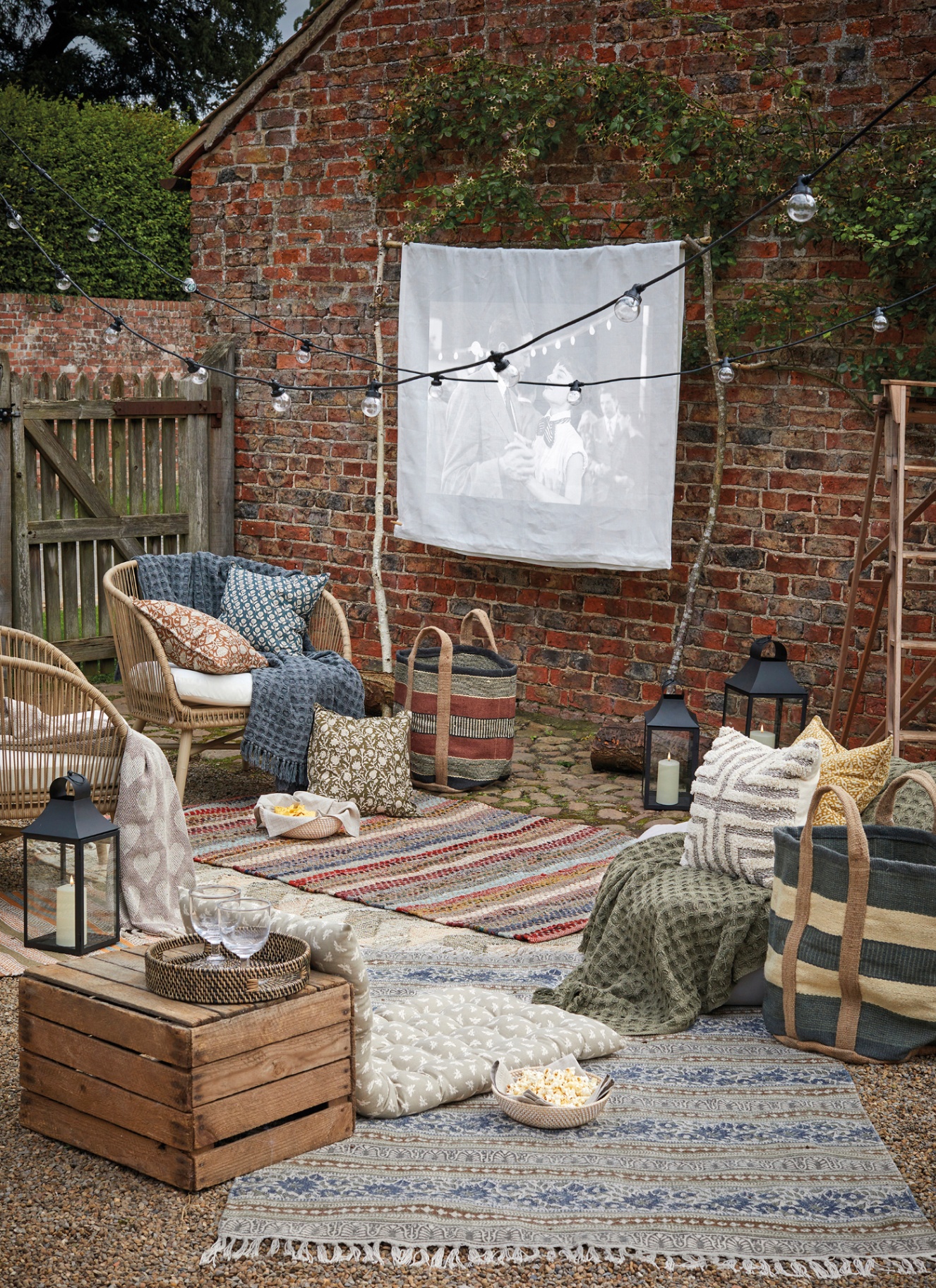 a garden with a red brick wall and a white sheet hanging on it with a movie being prjected, a string of festoon lights, earthy patterned cushions, throws and rugs and chairs facing the screen