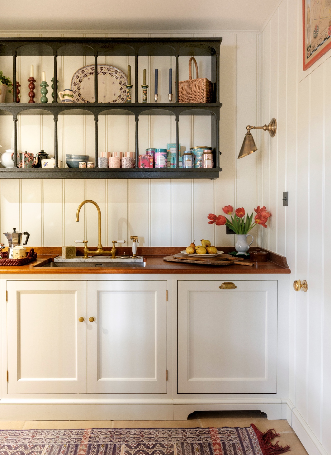 a kitchen sink area with white cabinets and white panelled walls and a large open shelving unit with places, candles and jars