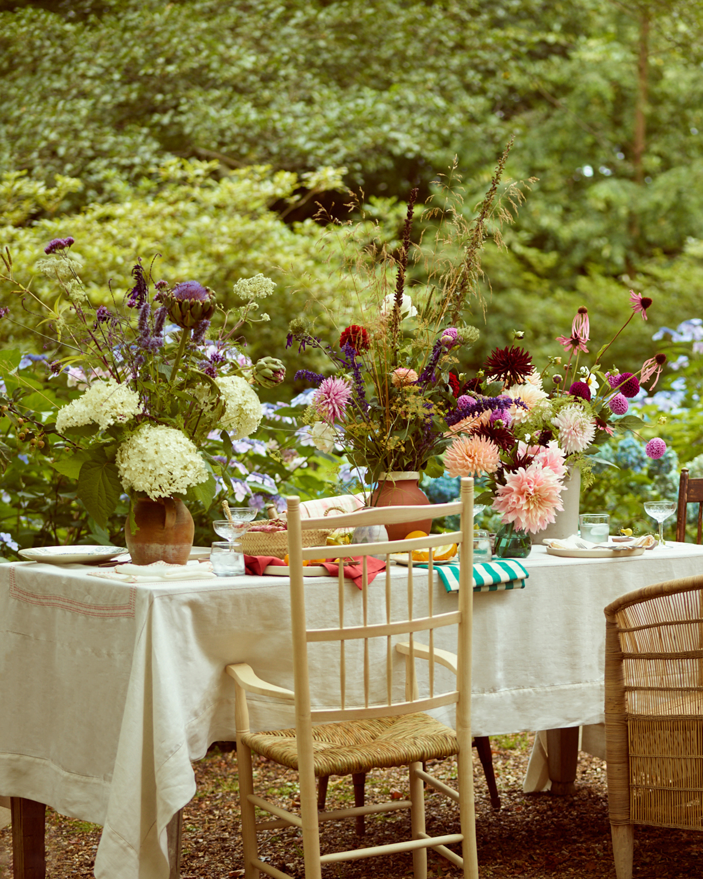 an outdoor table with a white table cloth, rattan dining chairs and multiple different vases filled with colourful summer flowers